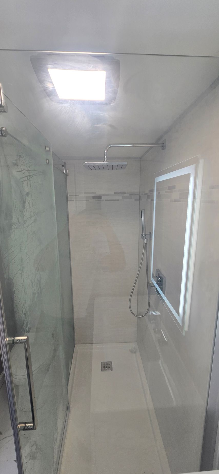 Narrow tiled shower with glass enclosure, overhead light, and white walls in a modern bathroom. By First Choice Bathroom Remodeling done in Norfolk, CT.