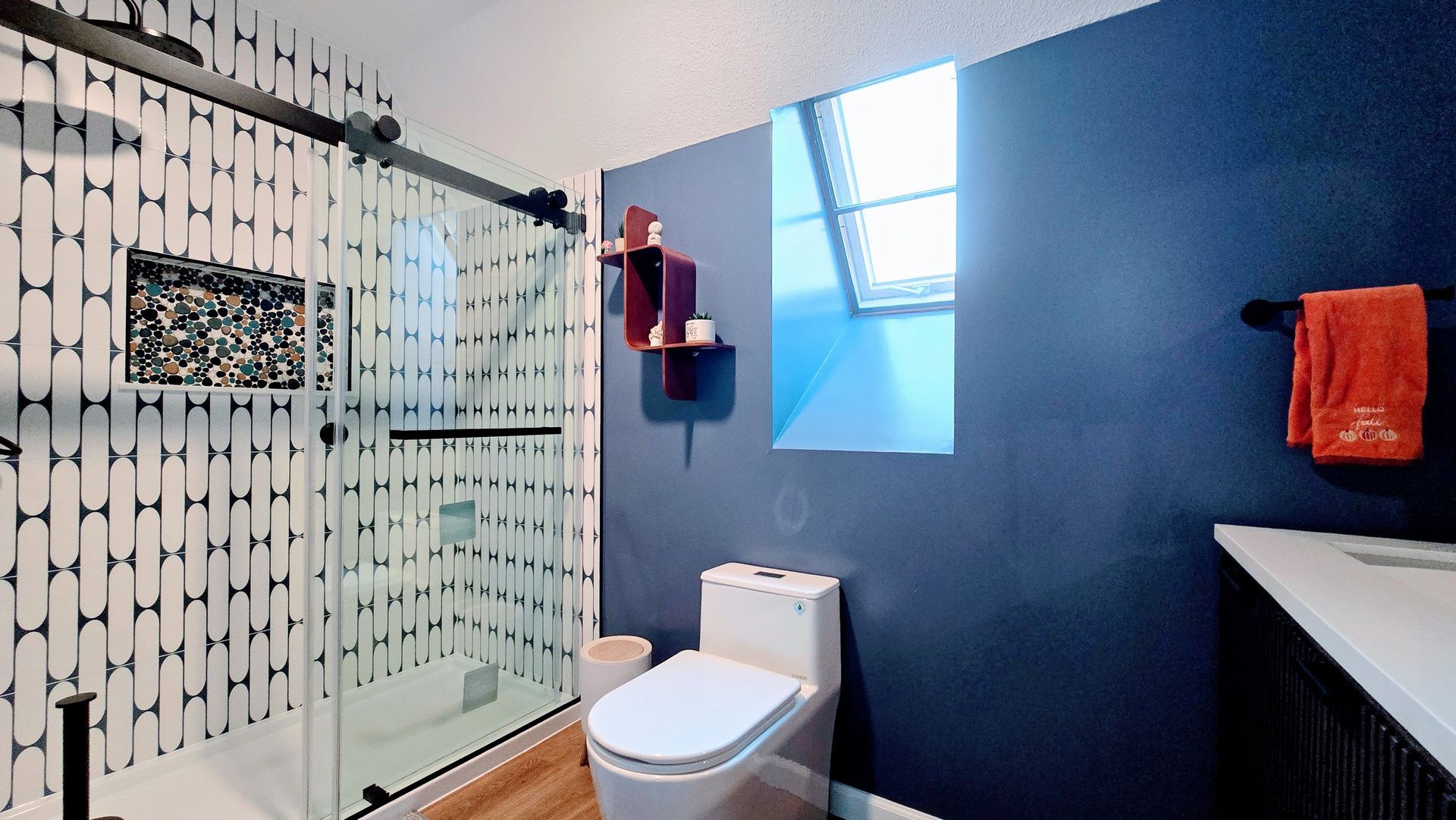 Modern bathroom with glass shower, white toilet, blue wall, skylight, and orange towels. By First Choice Bathroom Remodeling done in Bristol, CT.