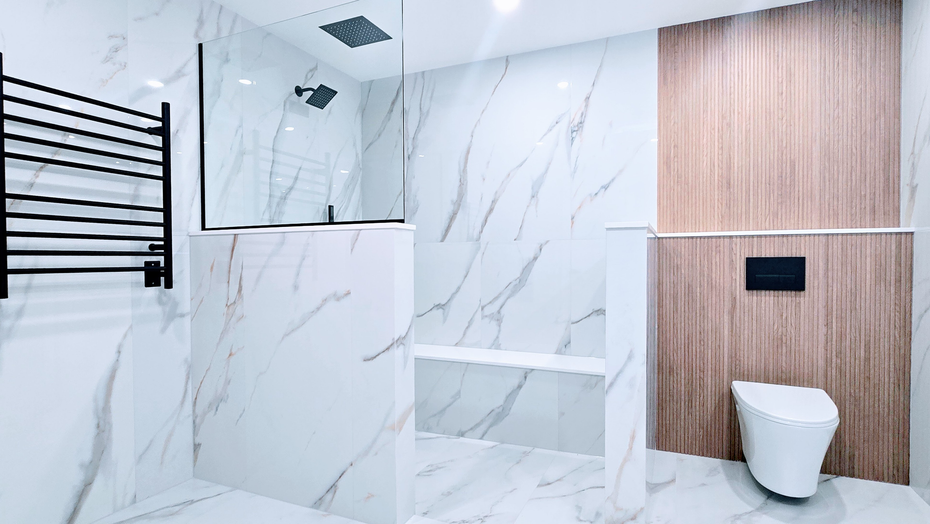Modern bathroom remodel in Southington, CT with white marble walls, wood accent panel, black towel rack, and white toilet