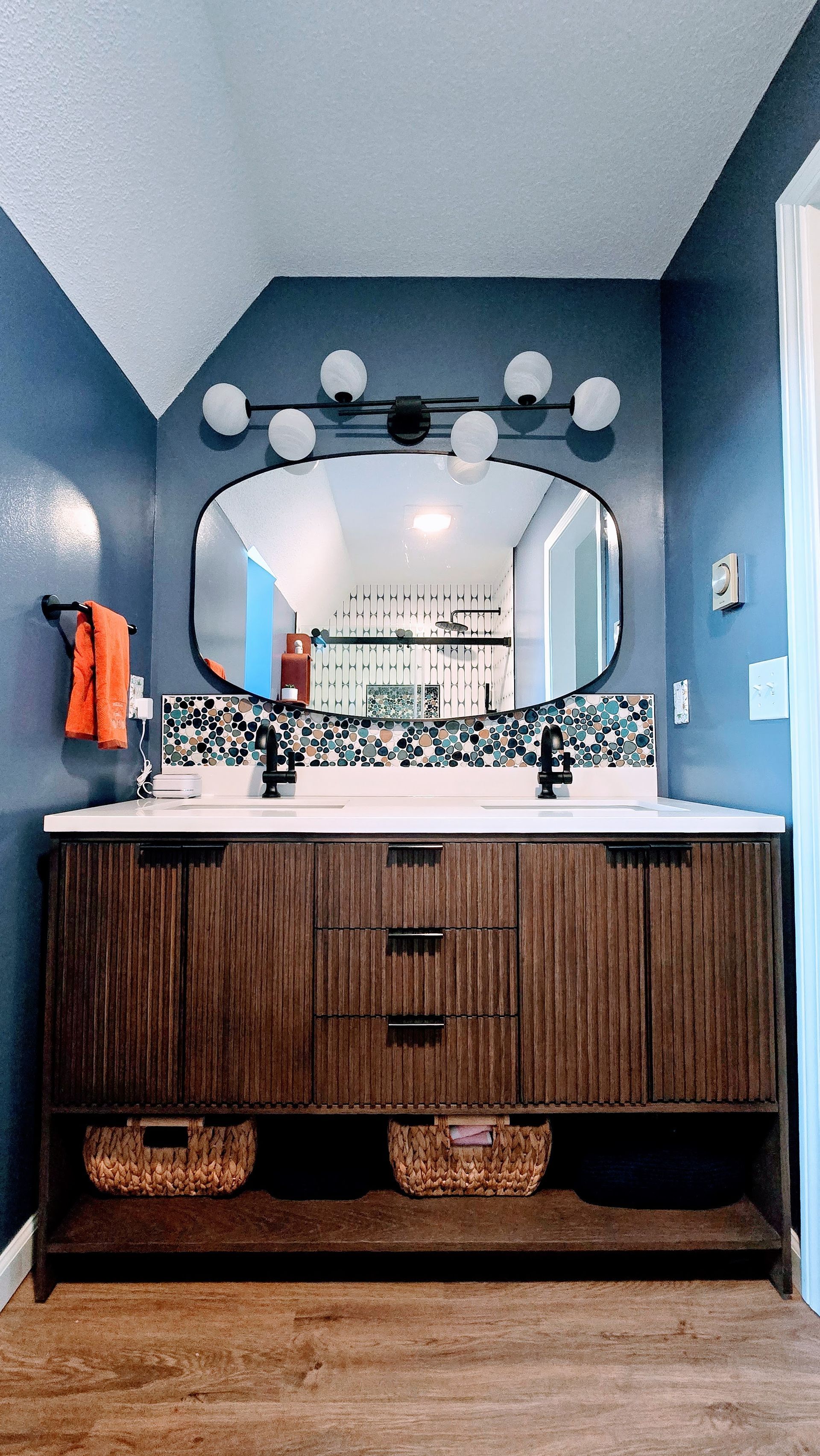 Blue bathroom vanity with oval mirror, wall sconces, white countertop, and woven baskets below. By First Choice Bathroom Remodeling done in Bristol, CT.