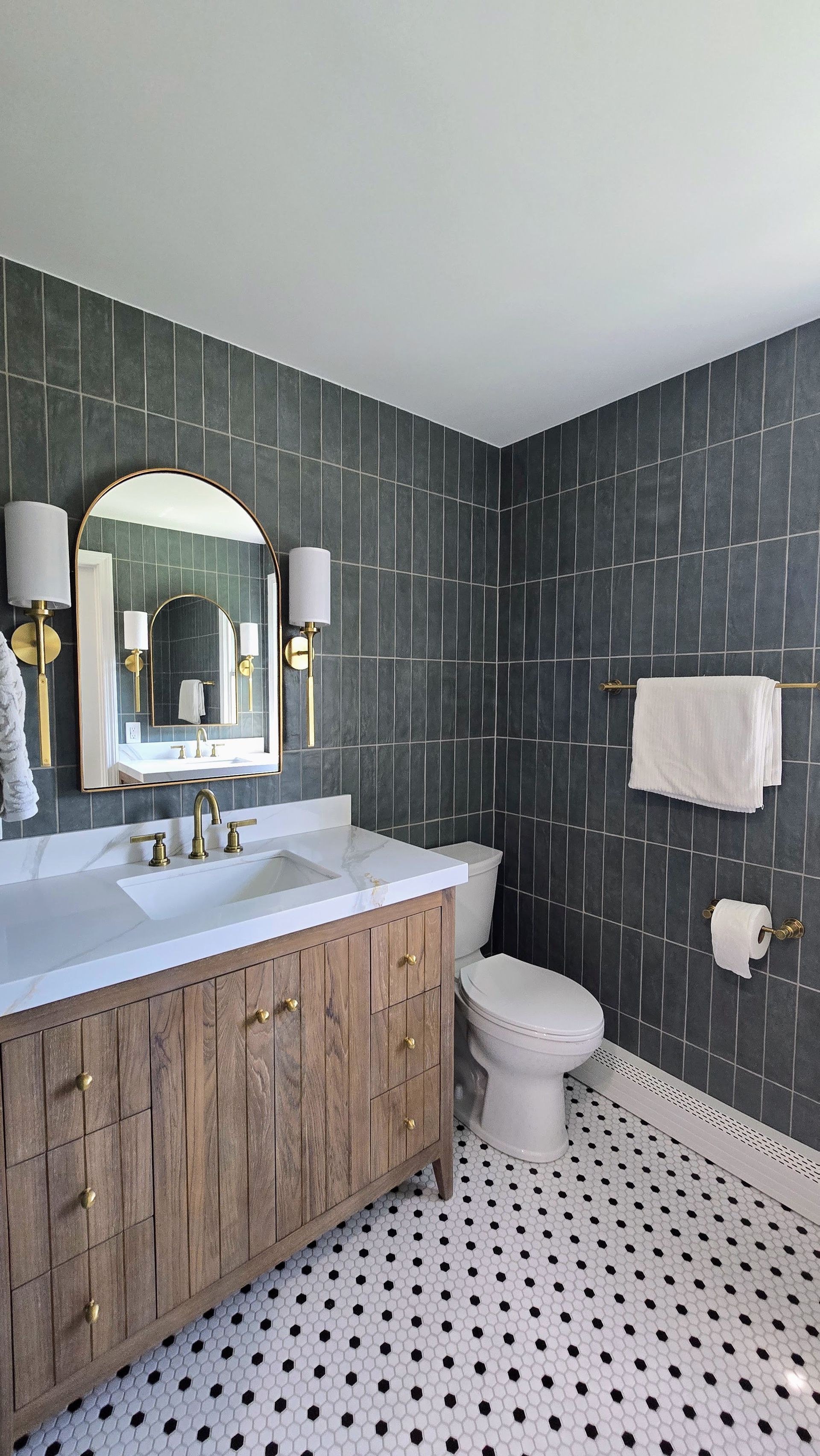 Small bathroom with dark tile walls, wood vanity, white sink and toilet, and black-and-white floor tiles. By First Choice Bathroom Remodeling done in Glastonbury CT.