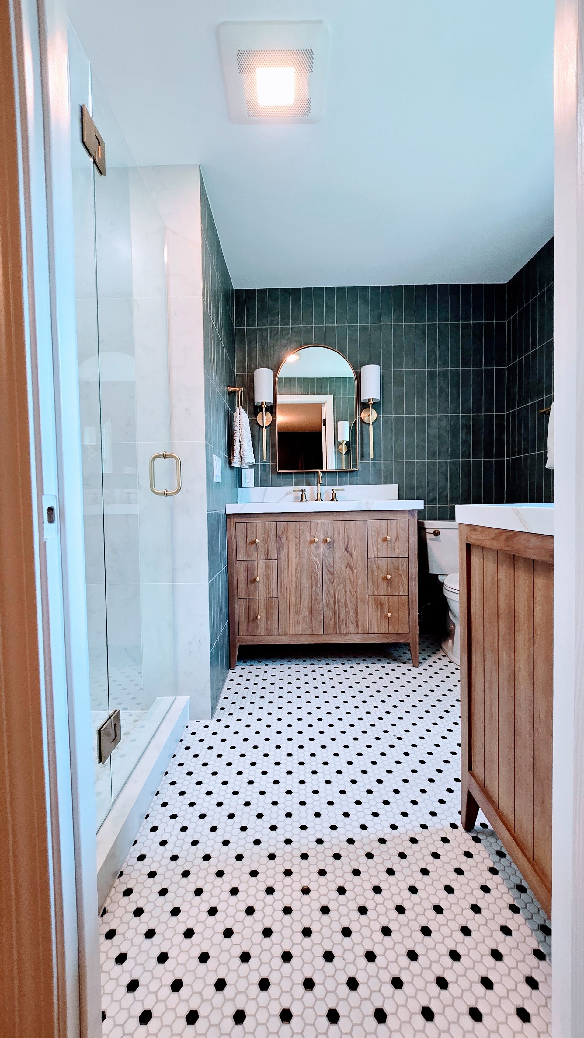Narrow bathroom with black tile walls, white polka-dot floor, wood vanity, mirror, and glass shower door. By First Choice Bathroom Remodeling done in Glastonbury CT.