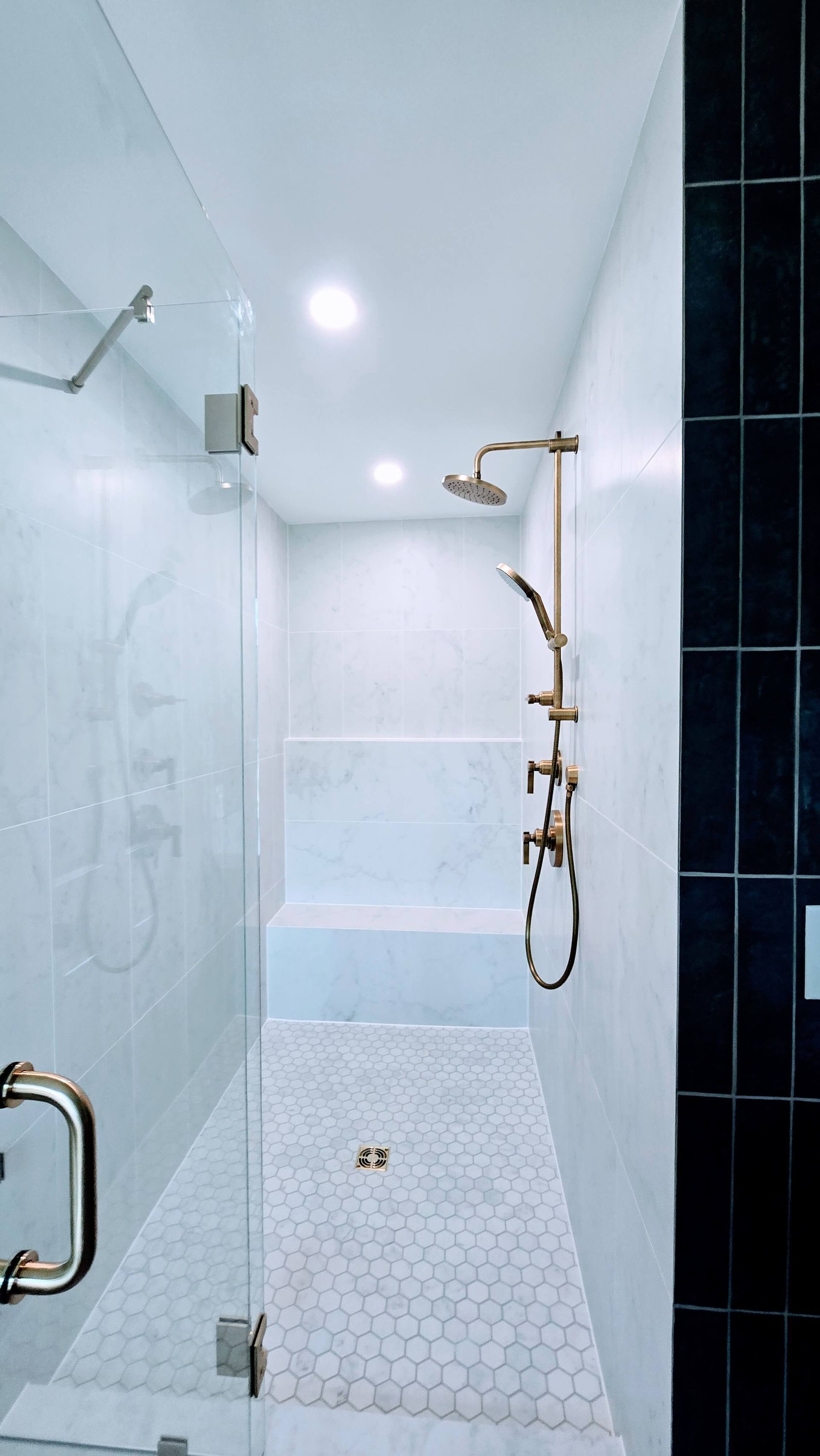 Modern tiled shower in Glastonbury, CT with glass door, white walls, black accent tile, and gold fixtures