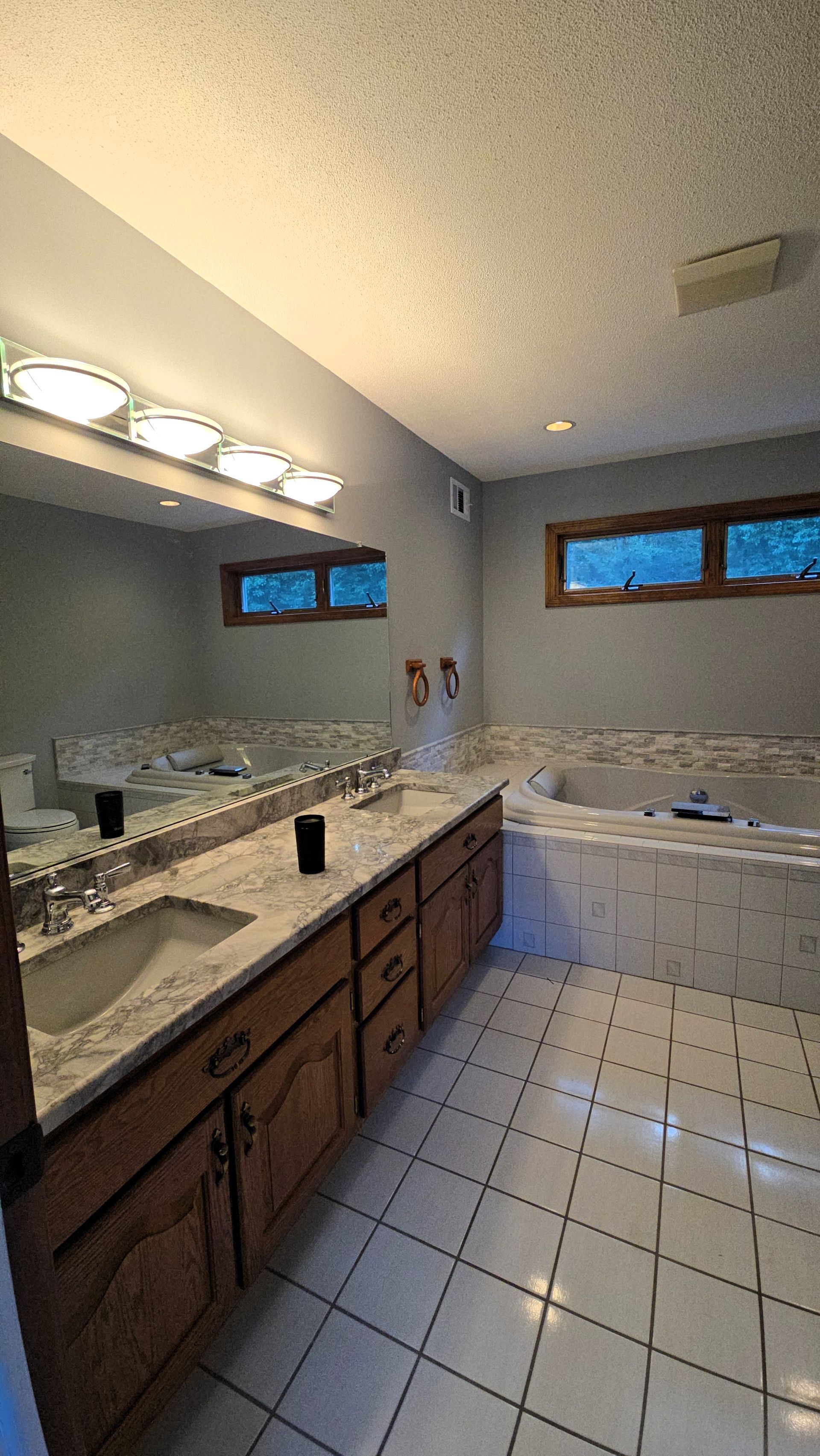Bathroom before renovation with double vanity, beige tile floor, wall mirror lights, and a built-in bathtub under small windows