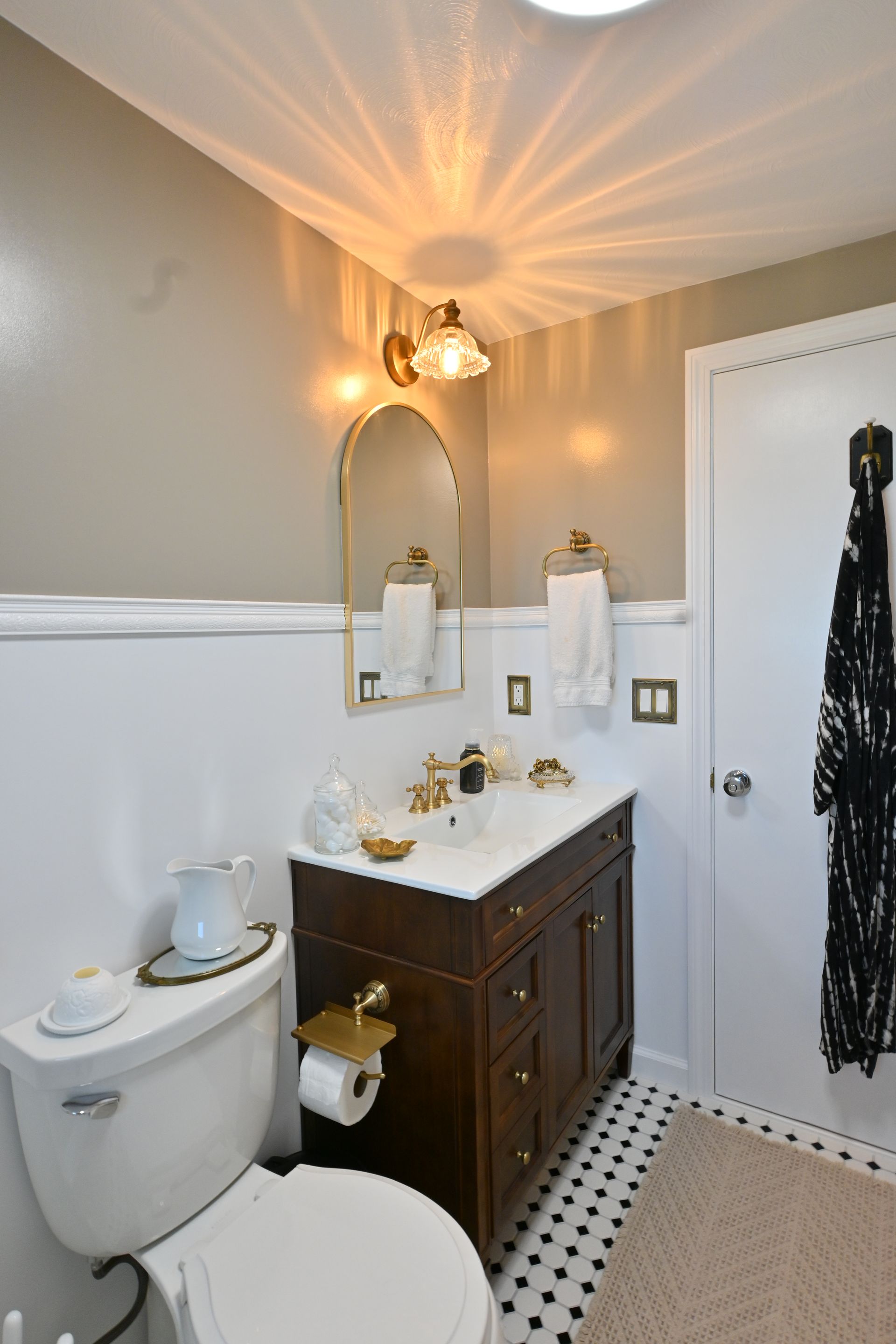 Small bathroom with white toilet, dark vanity, mirror, sconce, and black-and-white tile floor.