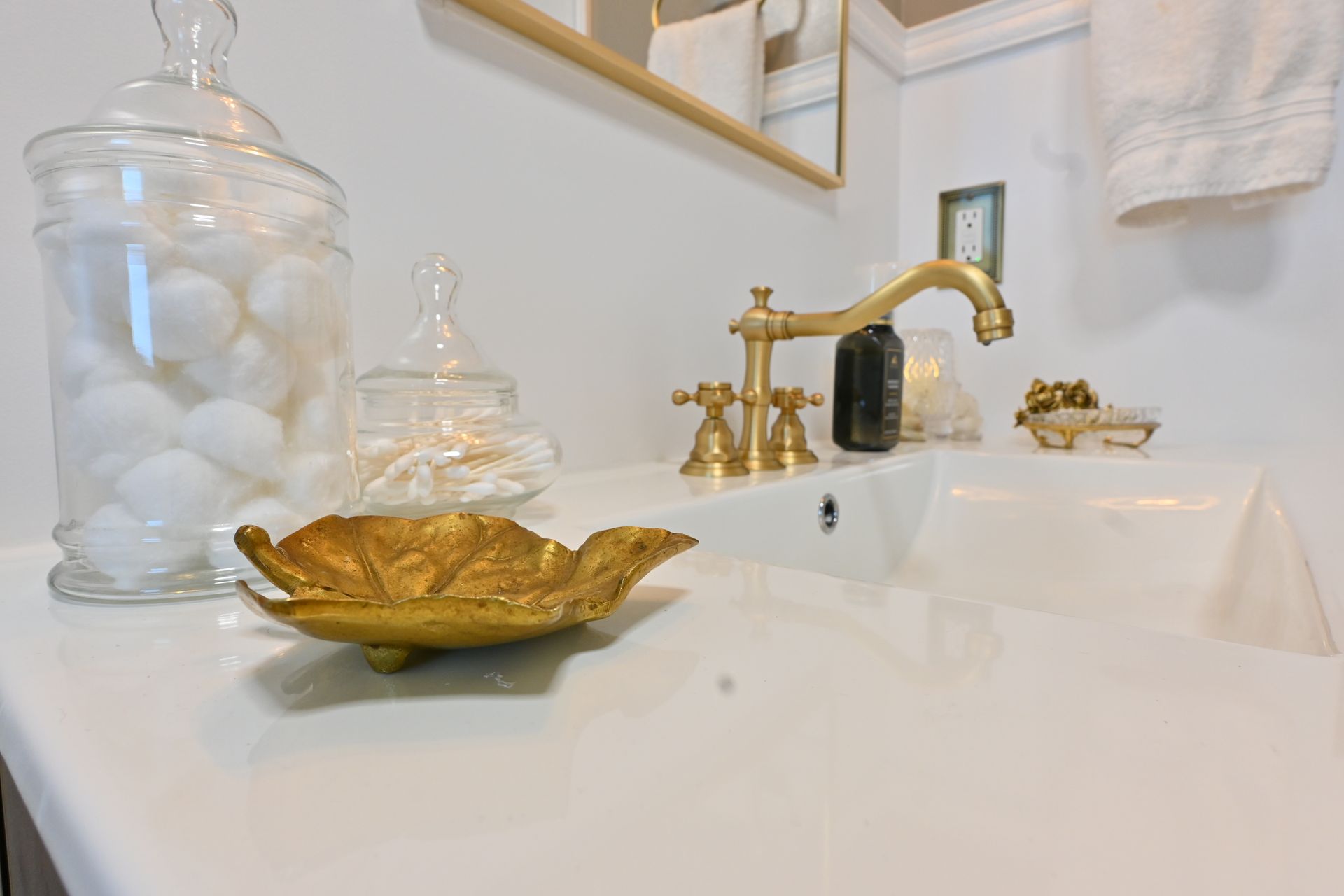Bathroom vanity with gold faucet, white sink, glass jars, and gold leaf dish on a white countertop