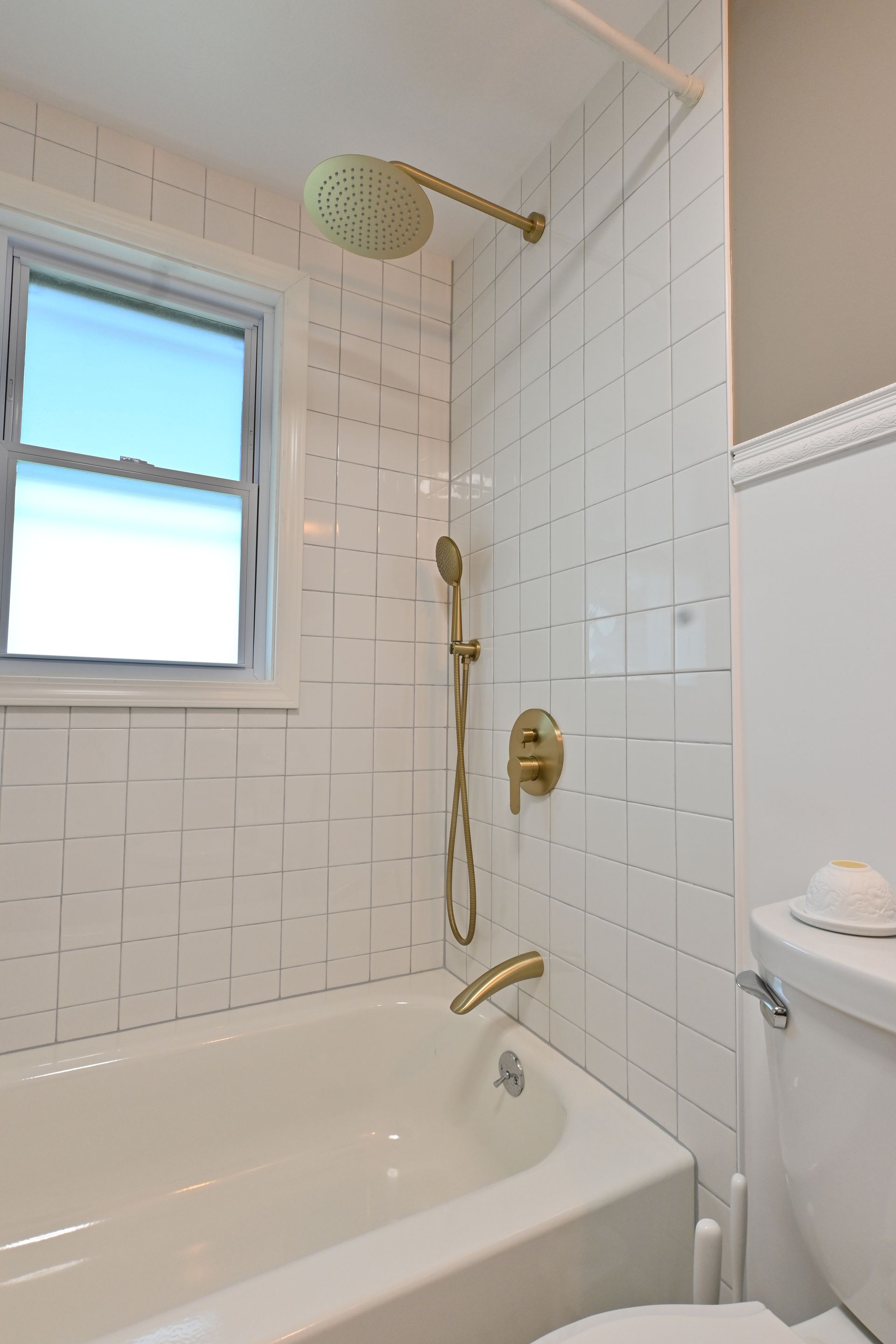 White tiled bathroom with bathtub, brass shower fixture, and window with blue sky visible