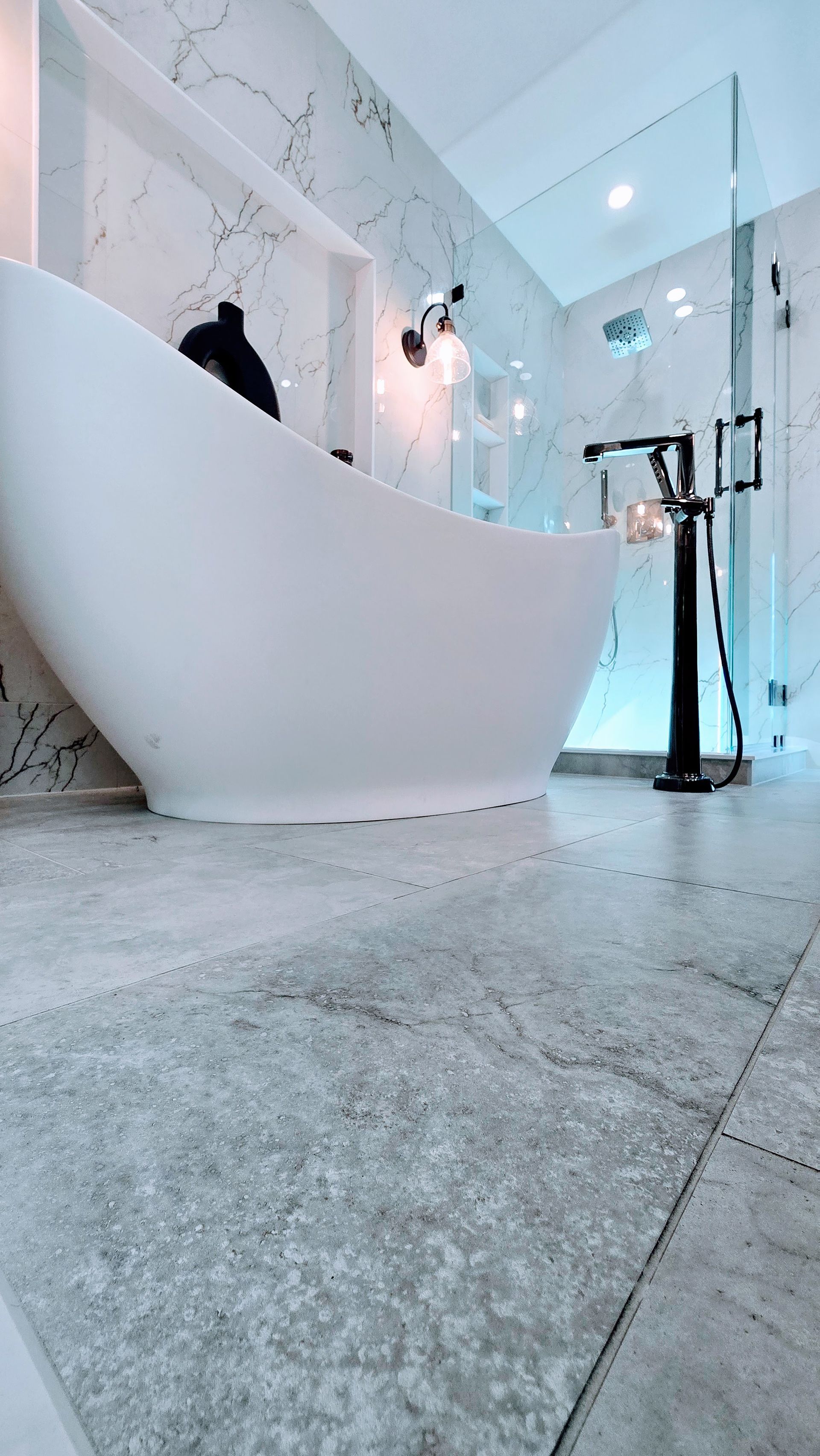 Modern white bathtub in Cromwell, CT in a bright bathroom with marble walls and gray floor tiles.
