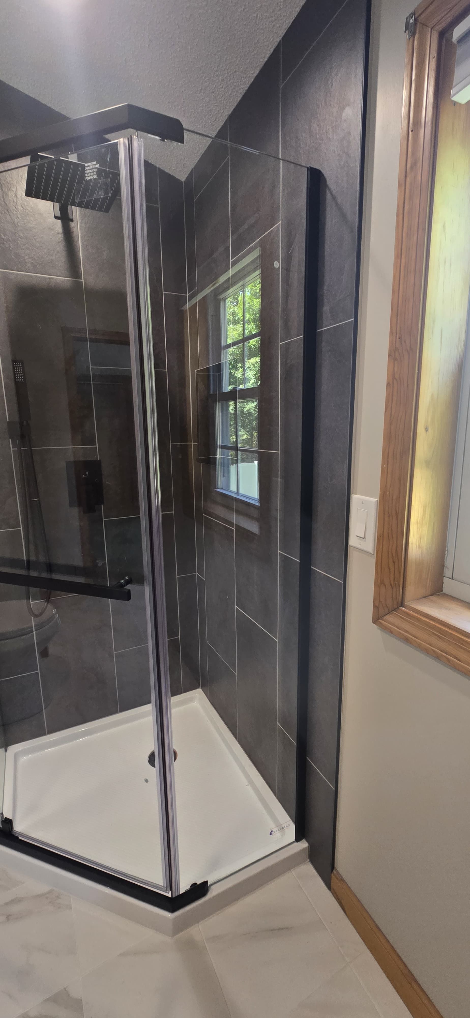 Glass shower enclosure with black trim in a bathroom, beside a wood-framed window. By First Choice Bathroom Remodeling done in Cheshire, CT.