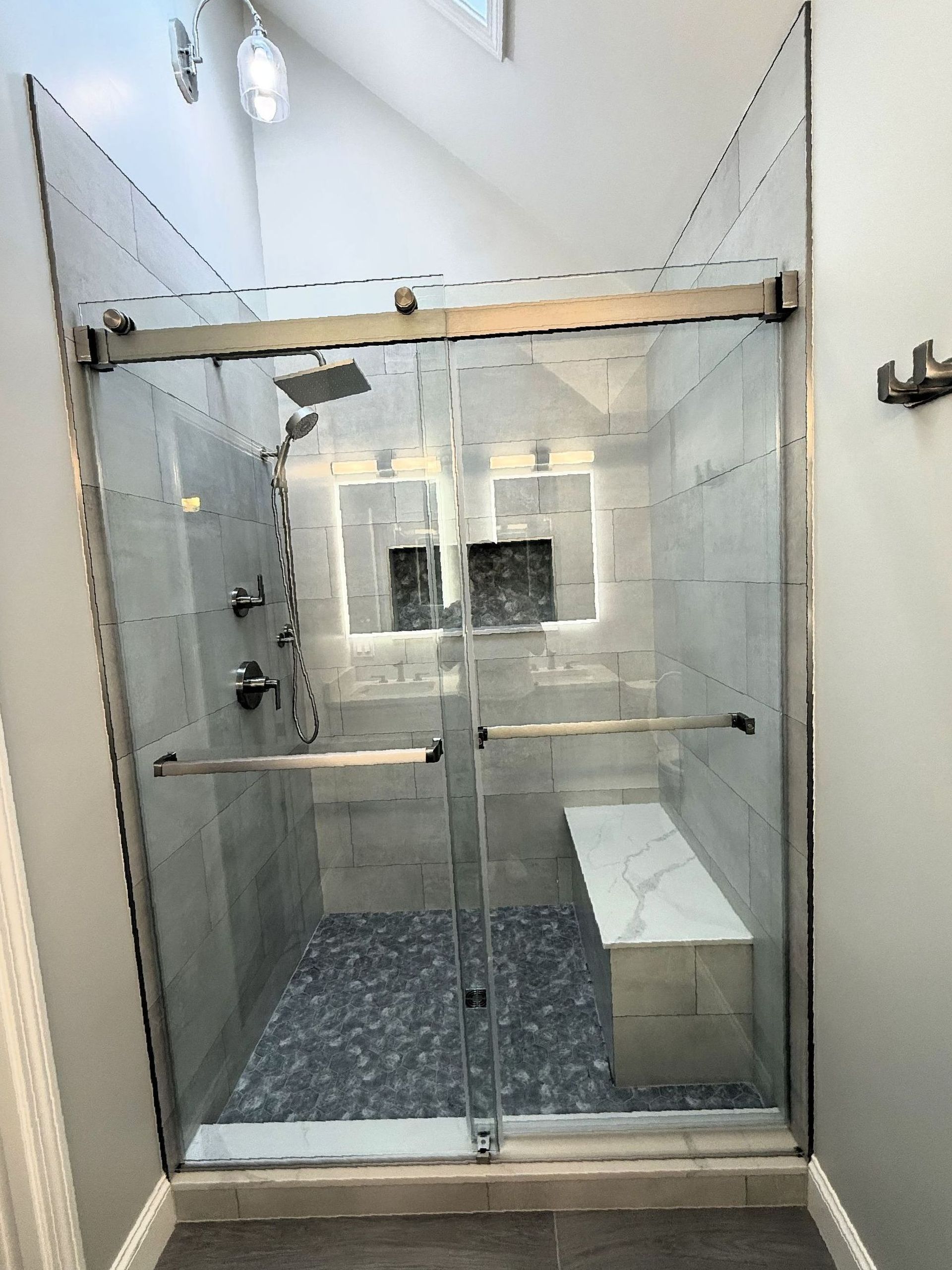 Glass-enclosed shower with gray tiled walls and a white bench, dark stone floor, and silver fixtures.