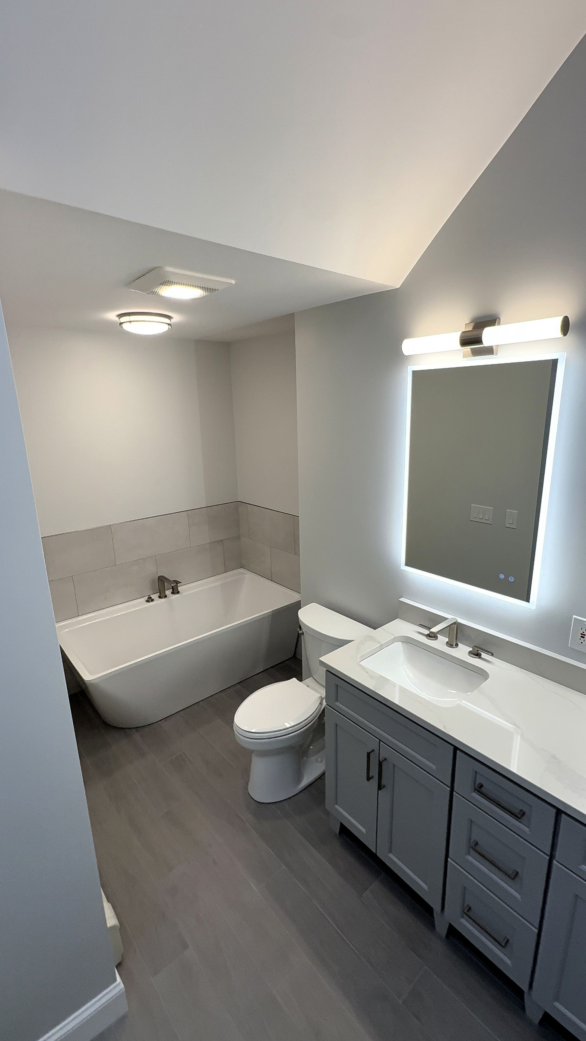 Modern bathroom with gray vanity, toilet, bathtub, and light-up mirror. Gray flooring, white walls.