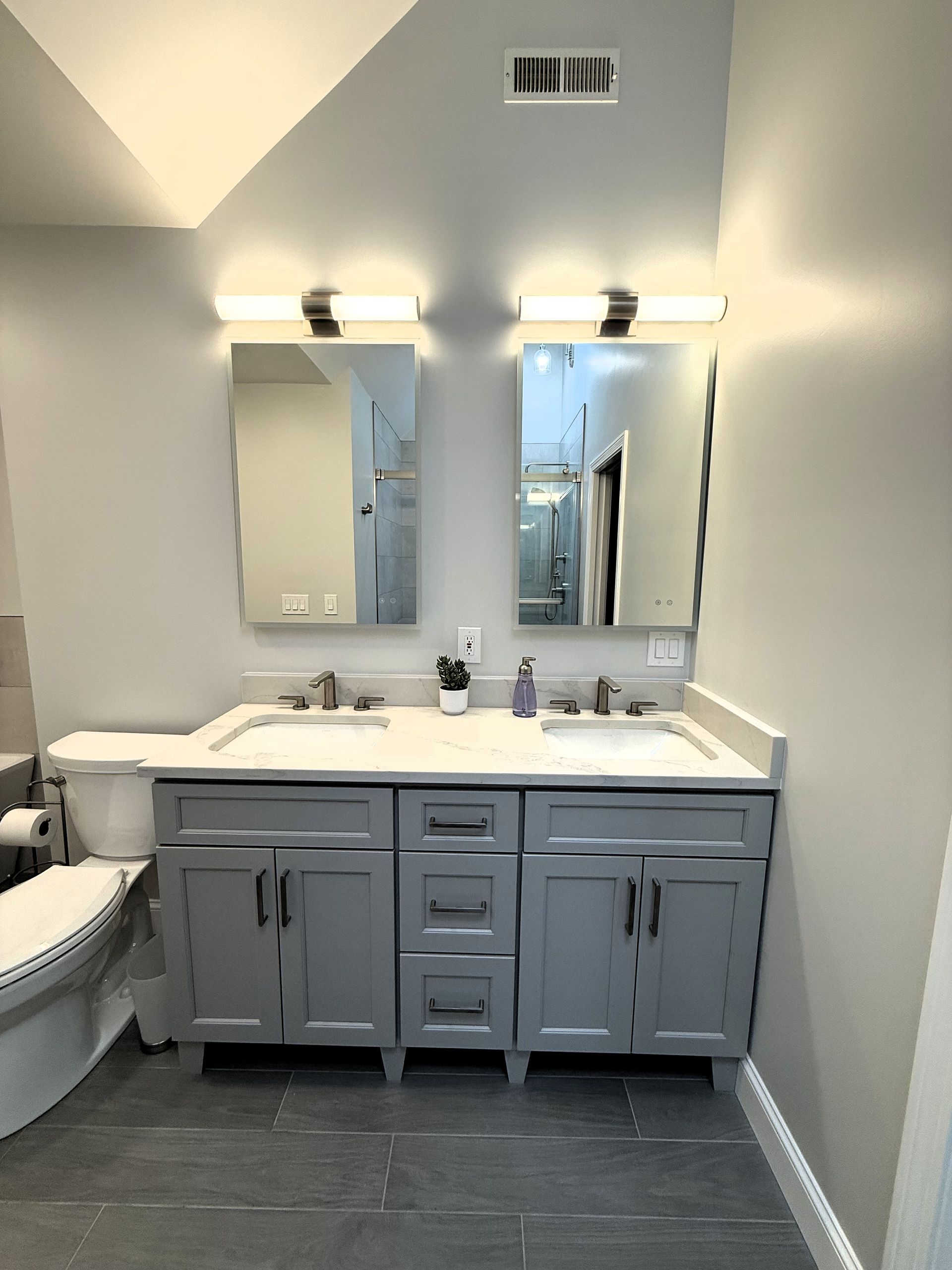 Gray bathroom with a double vanity, mirrors, lights, and a toilet.