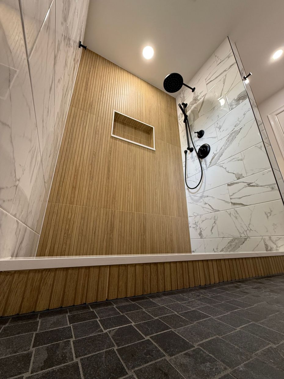 Modern walk-in shower in Berlin, CT with marble and wood-look tile, black fixtures, and a built-in shelf.
