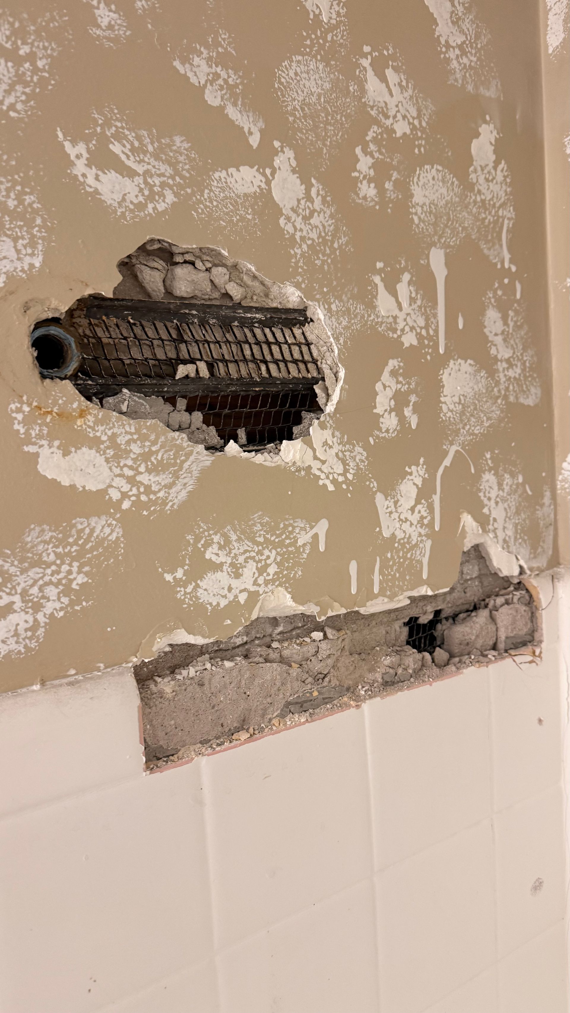 Damaged wall with exposed plumbing pipe and rough plaster repair above white tile