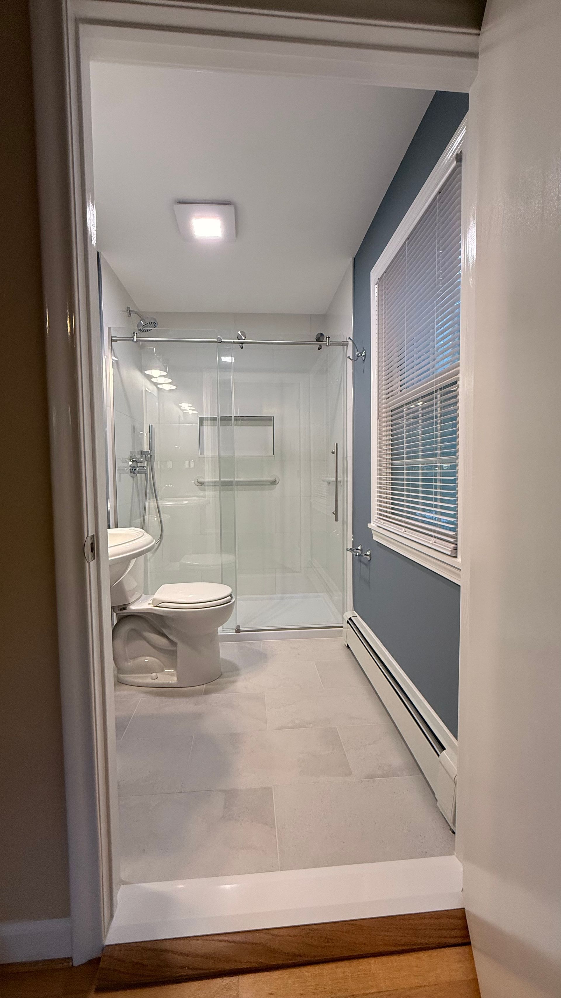 Modern bathroom with glass shower, toilet, and large window with blinds.