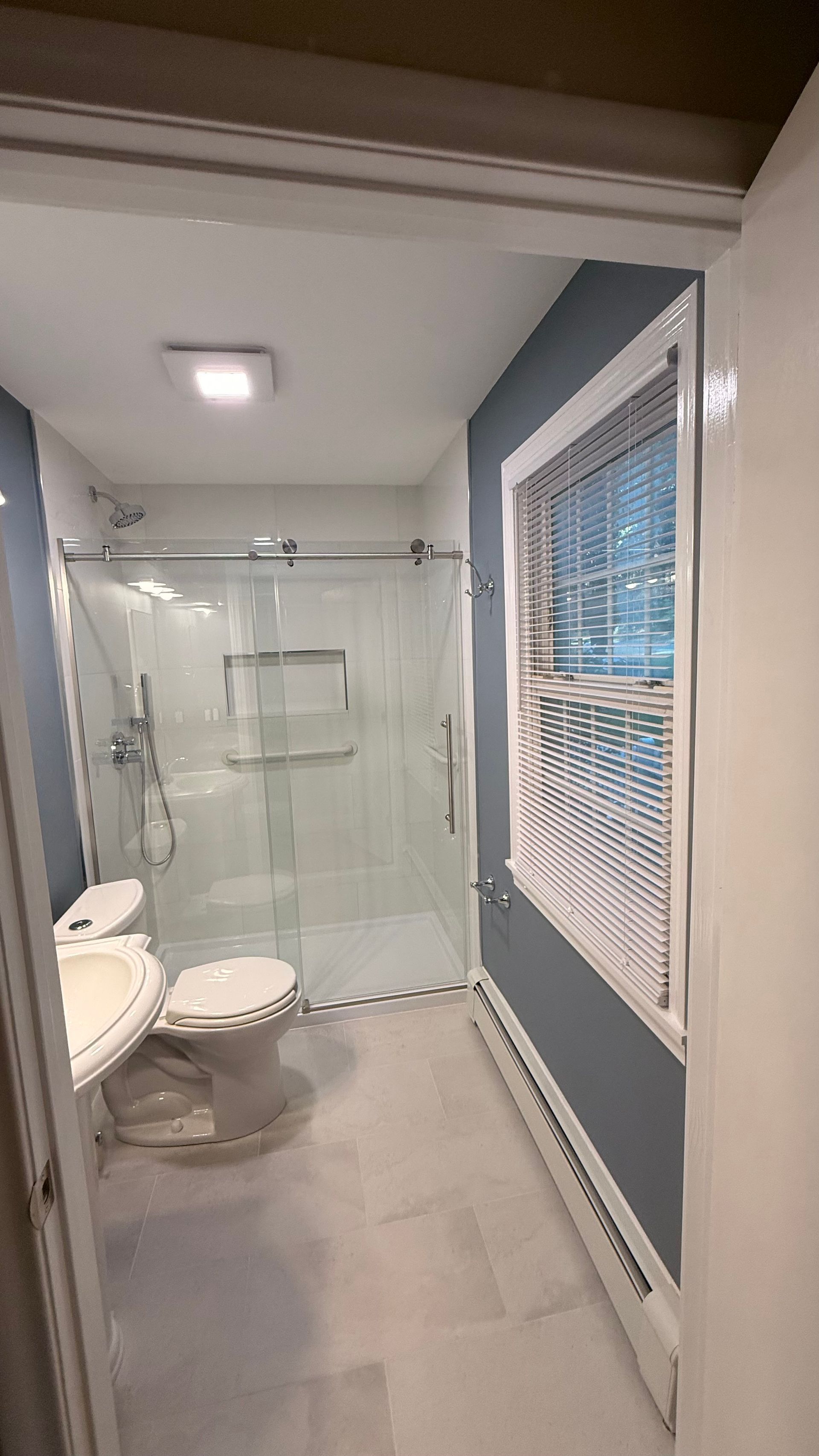 Bathroom with toilet, glass shower enclosure, and large window with blinds. By First Choice Bathroom Remodeling done in Avon, CT.