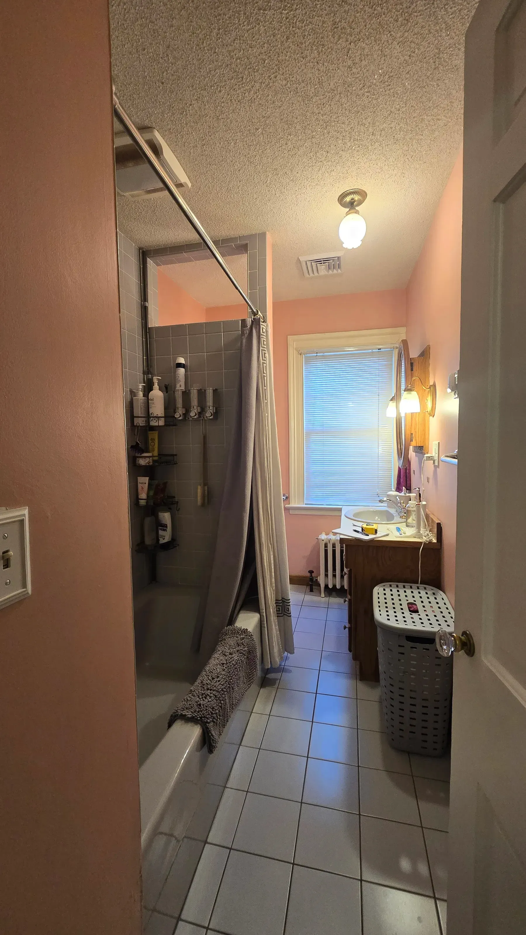 BEFORE Narrow hallway with white tiled floor, pink walls, a stairway, hanging light, and furniture at the end.