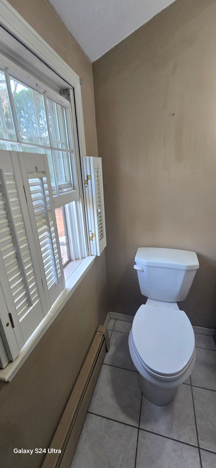 Toilet in bathroom next to window with shutters. Taupe walls, white toilet and tiles.