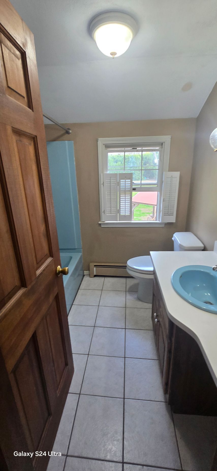 Bathroom with a brown door, blue sink, and a window. Light-colored walls and floor, white toilet, blue tub.