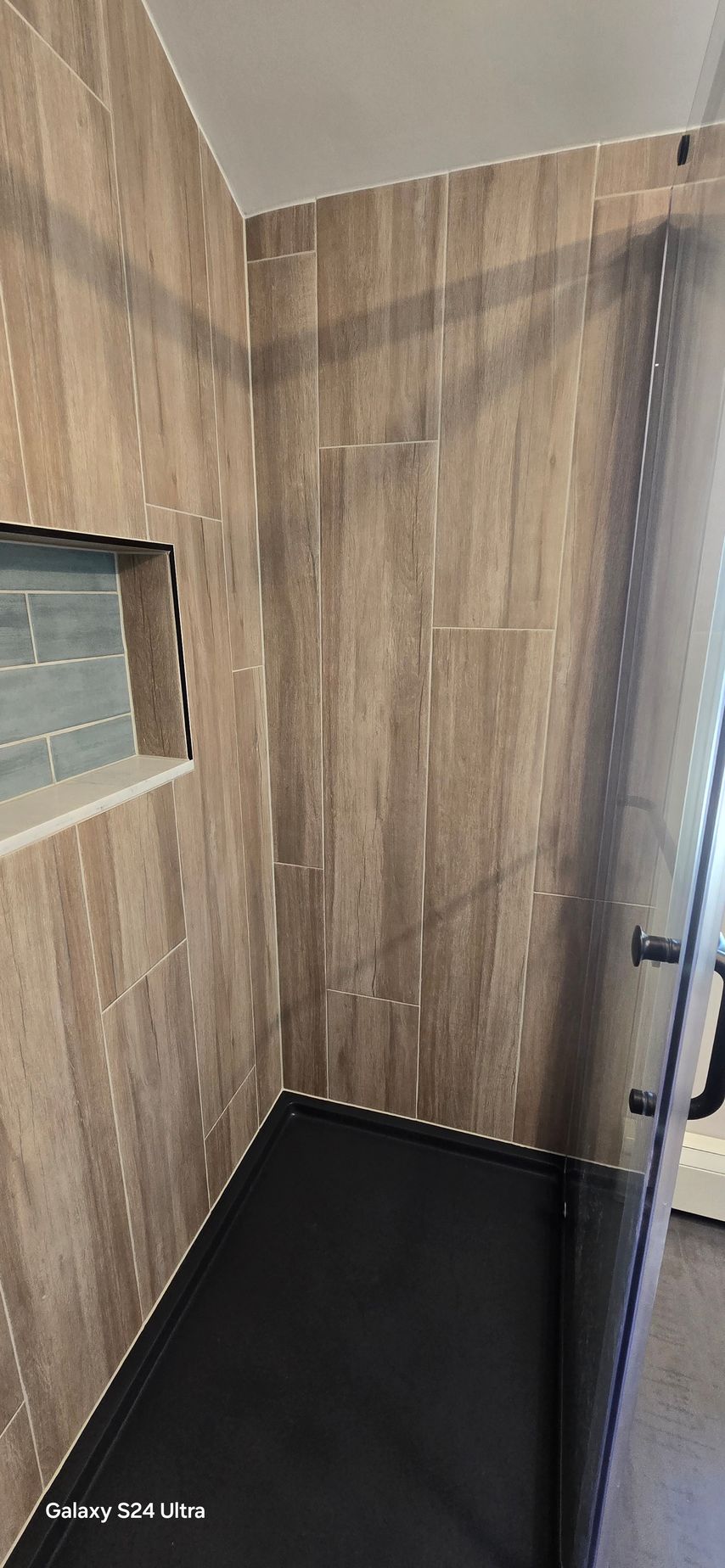 Shower interior with wood-look tile walls, black shower floor, and a glass door.