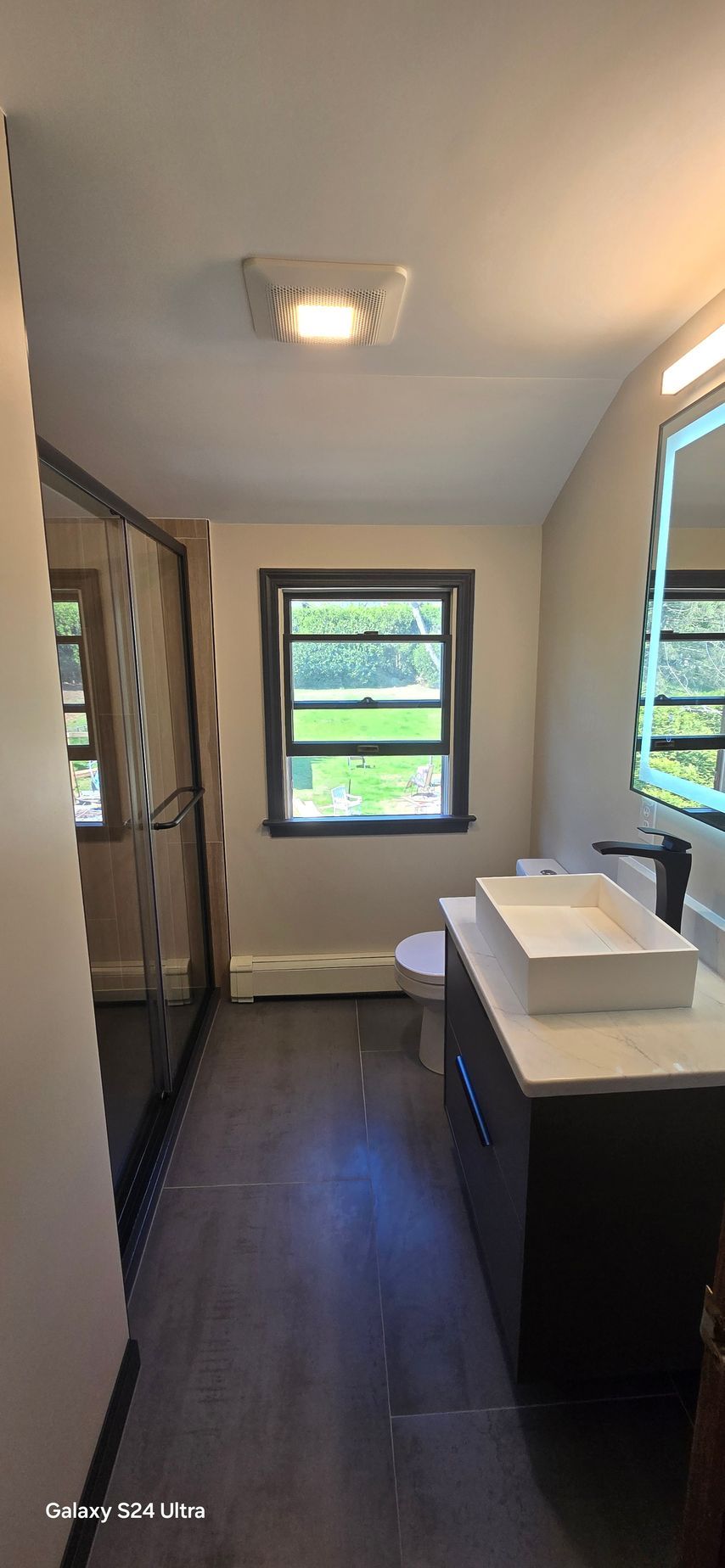 Bathroom with a dark vanity, white sink, window, and shower. Gray walls and wood-look flooring.