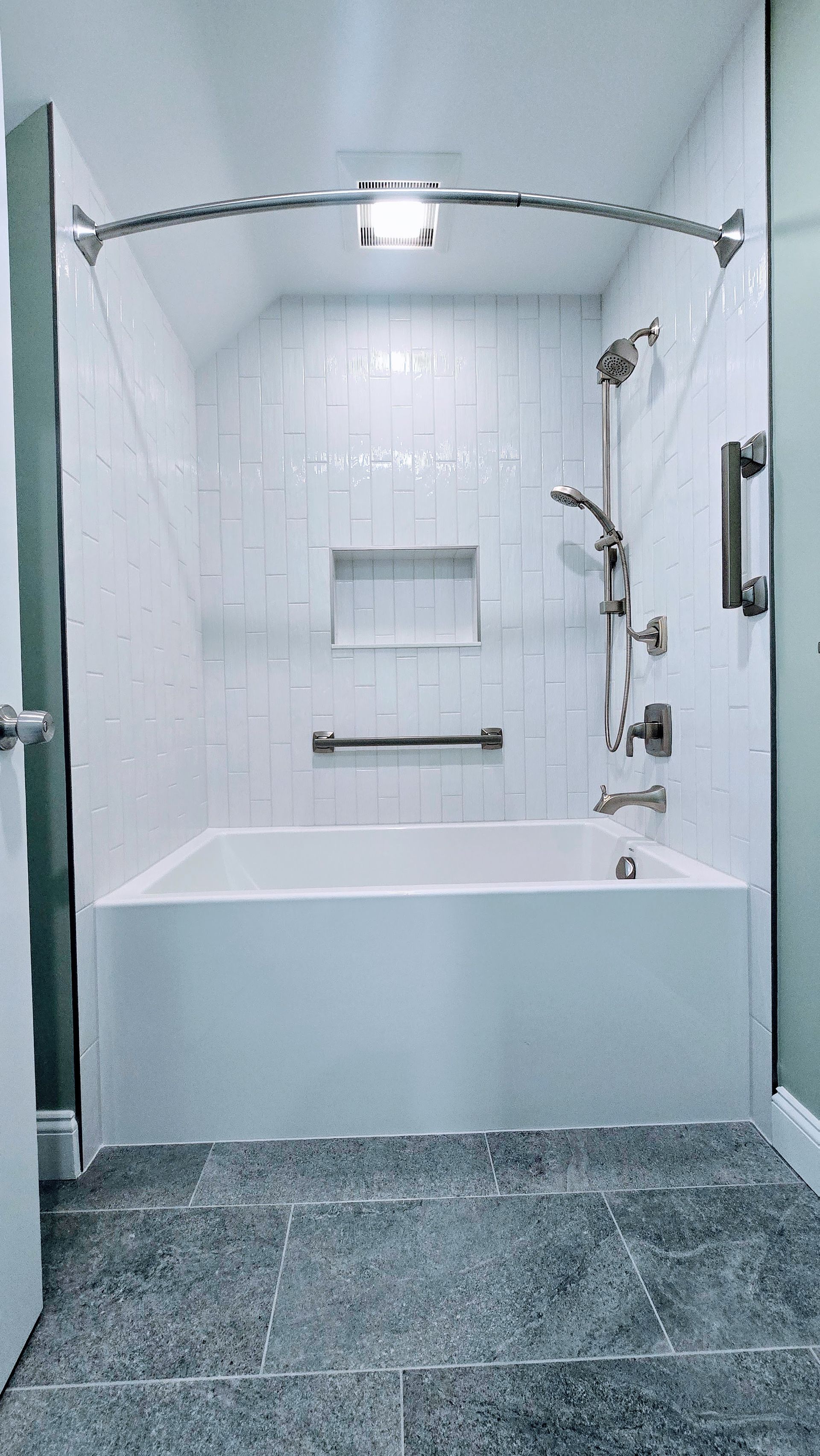 Tub with white-tiled accessible shower in Newington, CT with grab bars and a bathtub. Dark gray tiled floor.