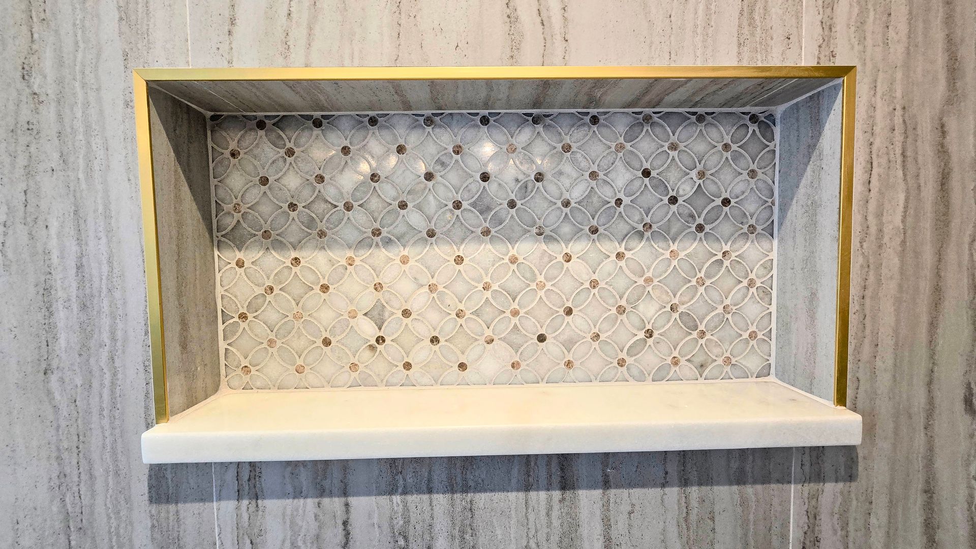 A rectangular shower niche in Cheshire, CT with a white marble shelf, a gold metal frame, and a decorative patterned tile back wall.