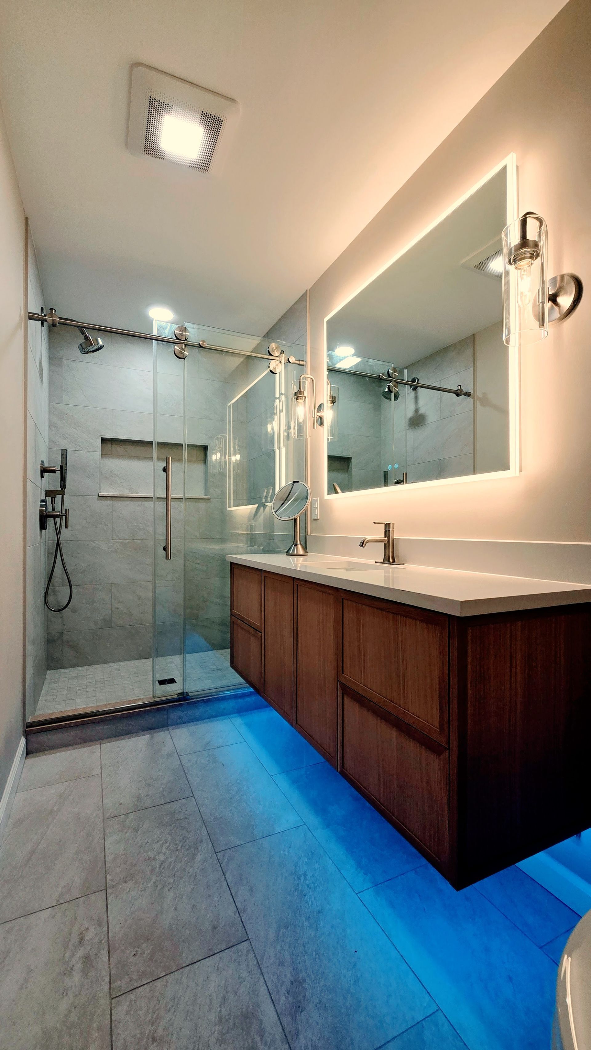 Modern bathroom in Simsbury, CT with a glass-enclosed shower, wood vanity, illuminated mirror, and blue under-cabinet ambient lighting.