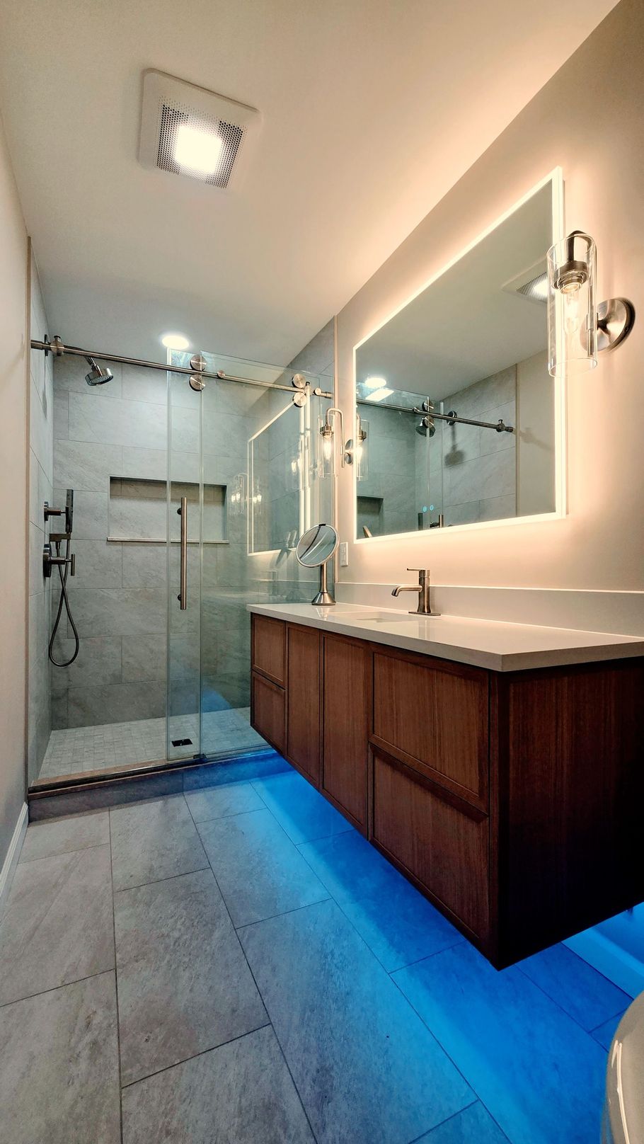 A modern bathroom in Simsbury, CTwith a wooden floating vanity, illuminated mirror, walk-in glass shower, and blue under-cabinet lighting.