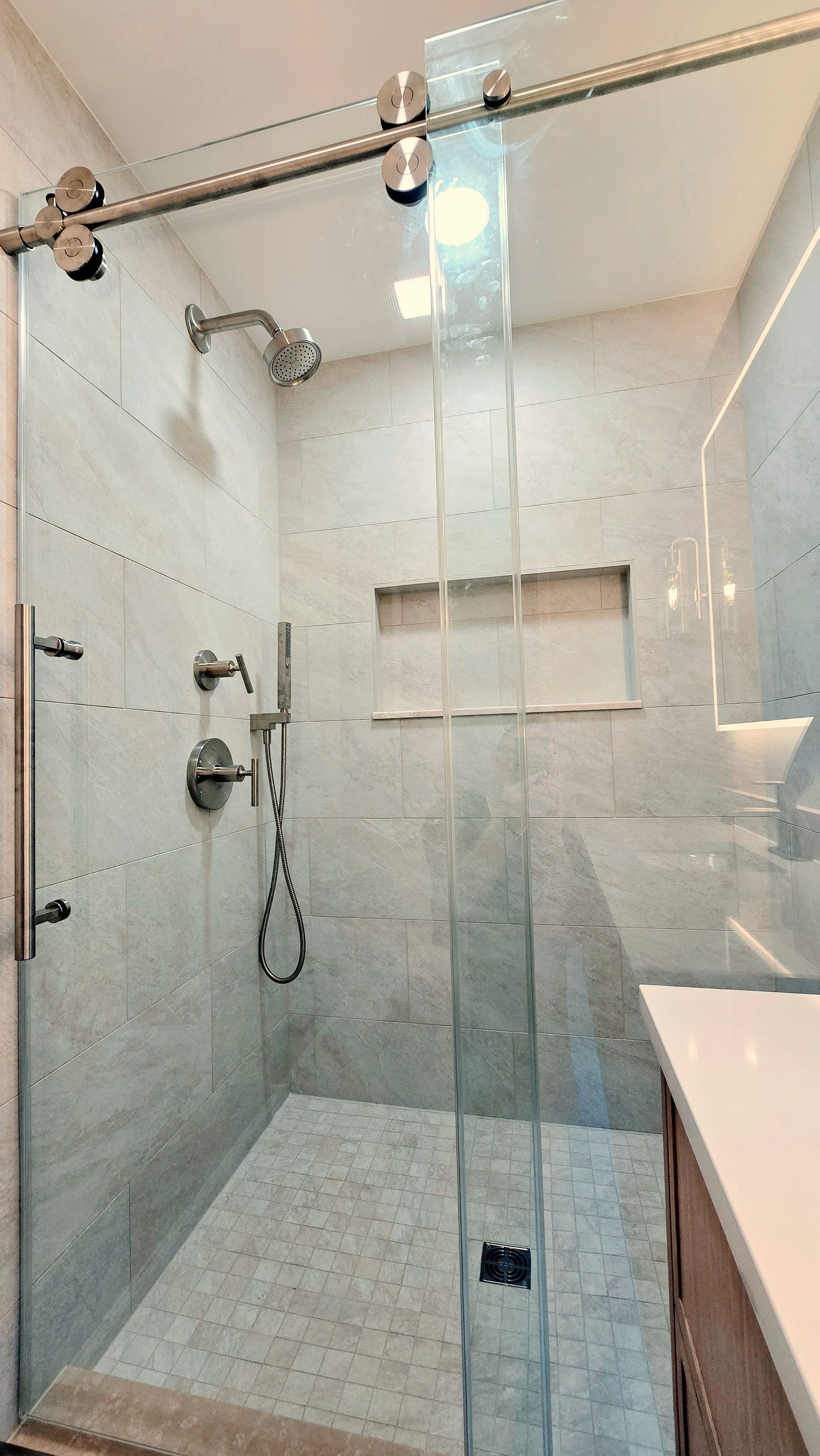 A modern shower in Simsbury, CT with light gray tiled walls, a sliding glass door, a rainfall showerhead, and a recessed wall niche.