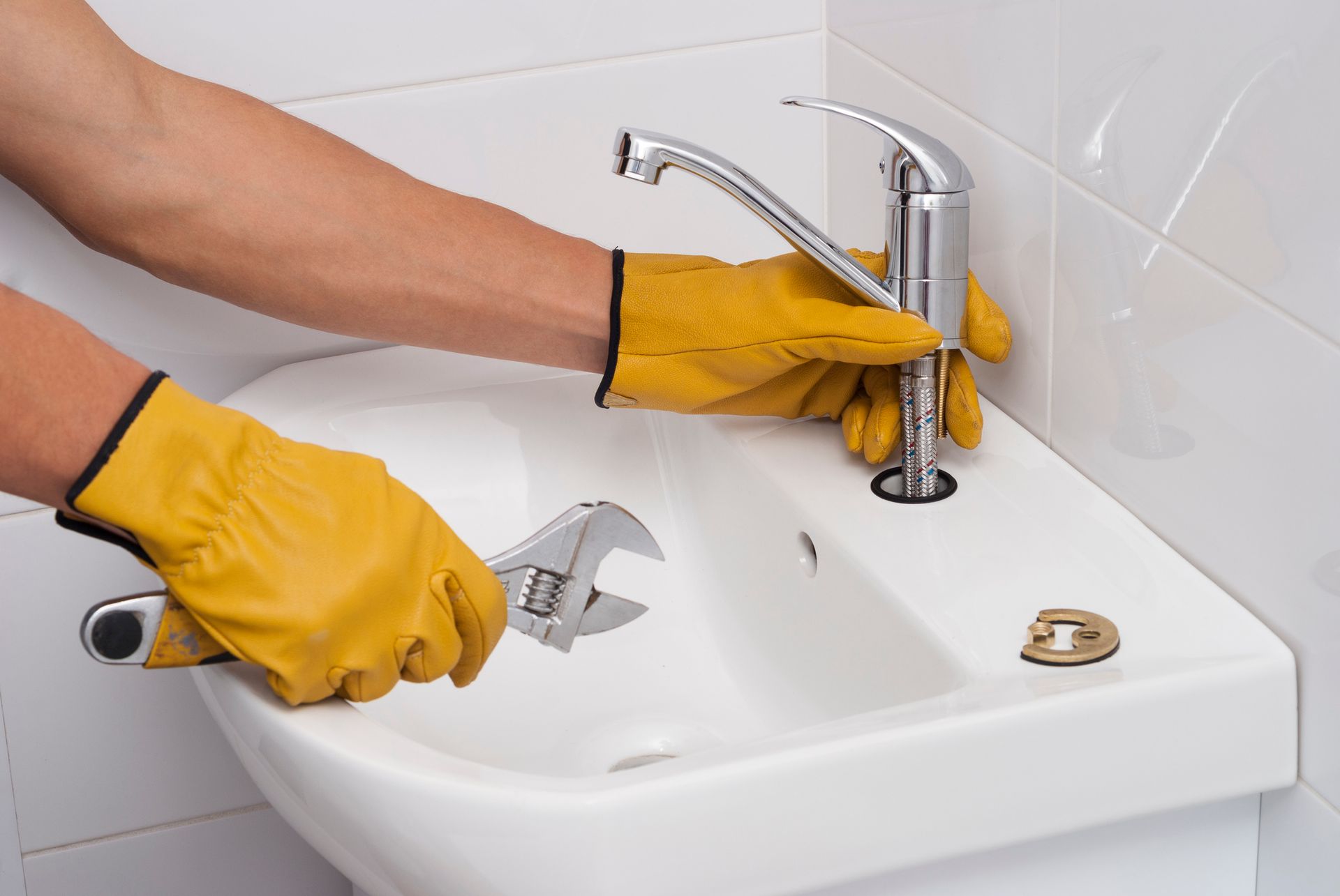 A plumber wearing yellow gloves installs a new silver faucet in a white bathroom sink.