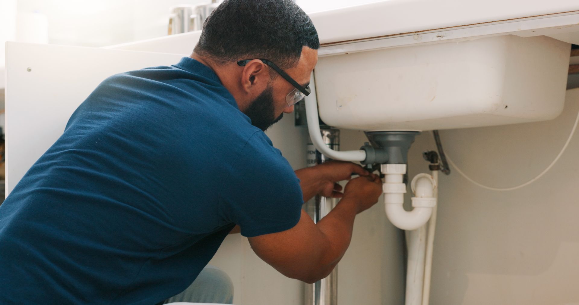 Plumber wearing safety glasses repairing pipes under bathroom sink.