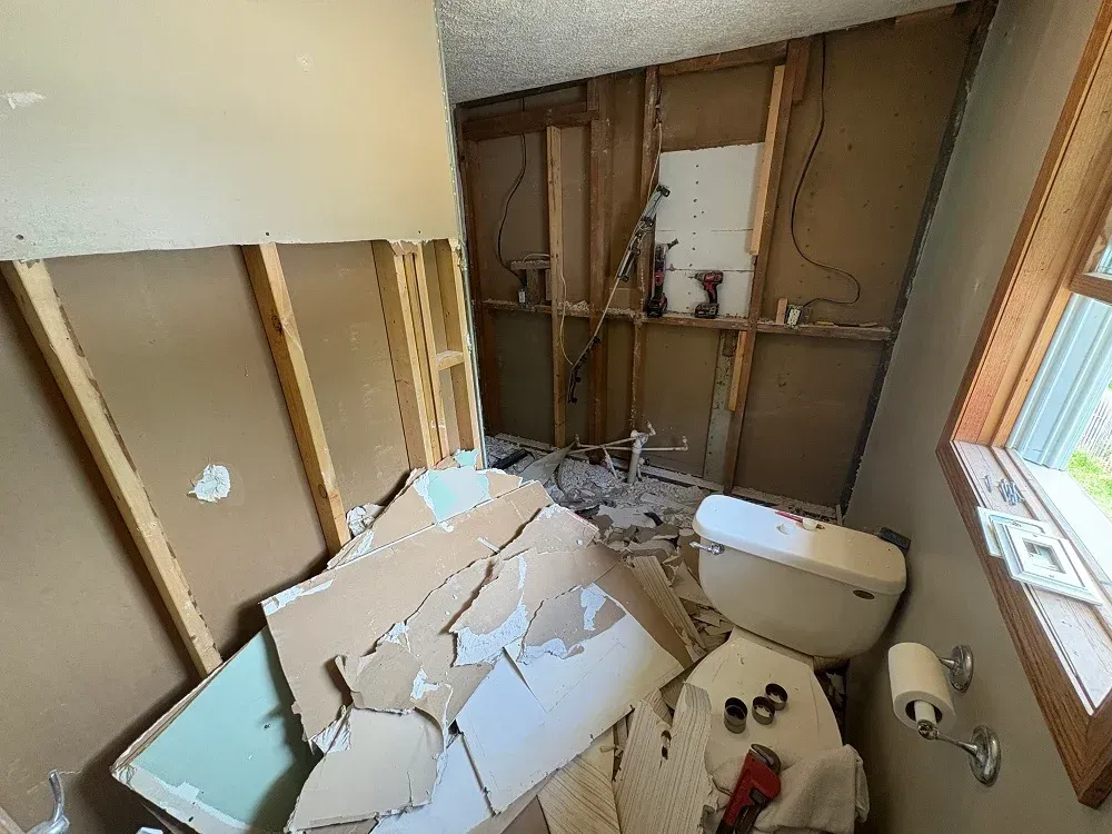 A bathroom undergoing demolition, with torn drywall on the floor and open wall studs exposing the plumbing and wiring.