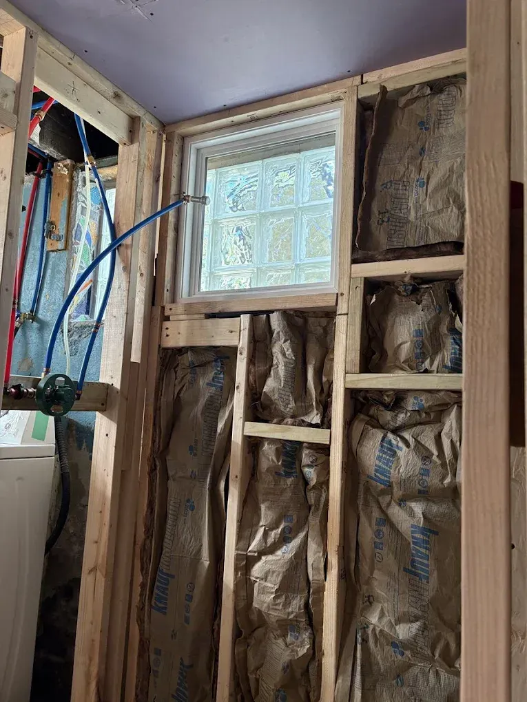 An interior view of wooden wall framing, insulation, a glass block window, and exposed blue and red plumbing pipes.