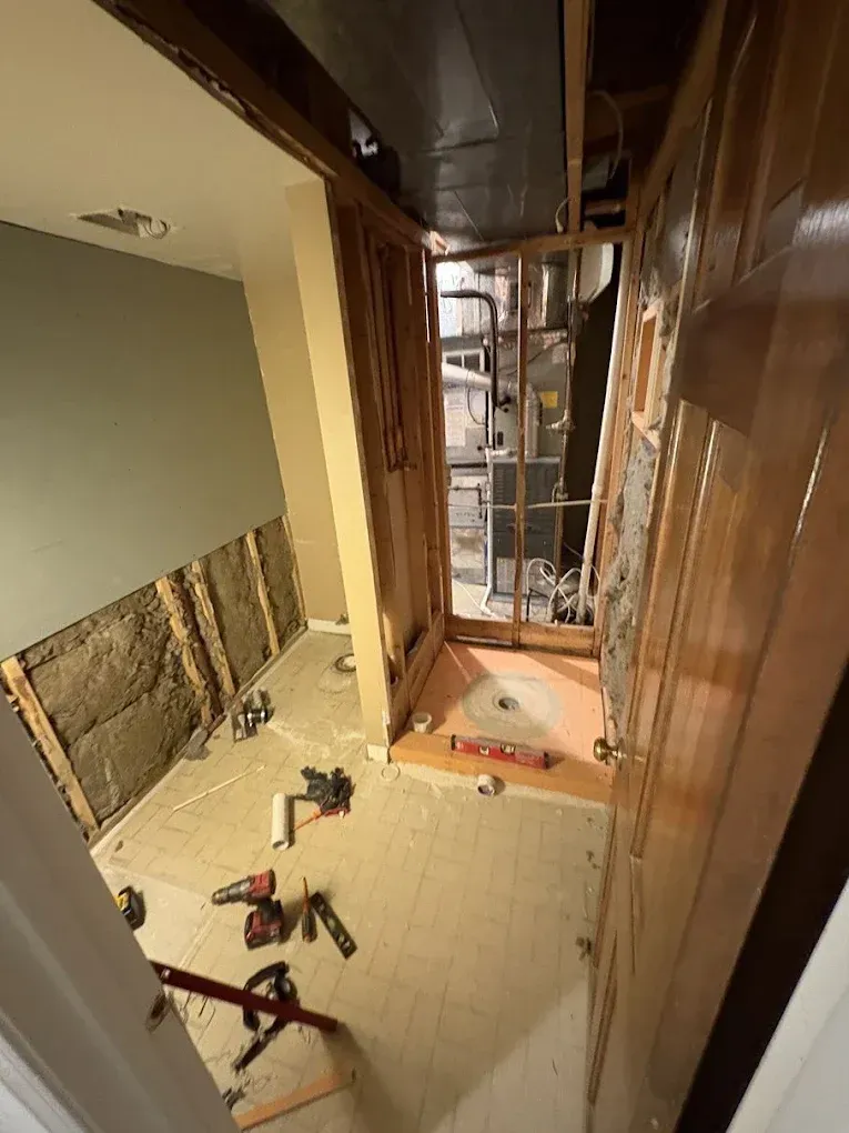 An unfinished room under renovation featuring exposed wood studs, insulation, a partially tiled shower floor, and tools.