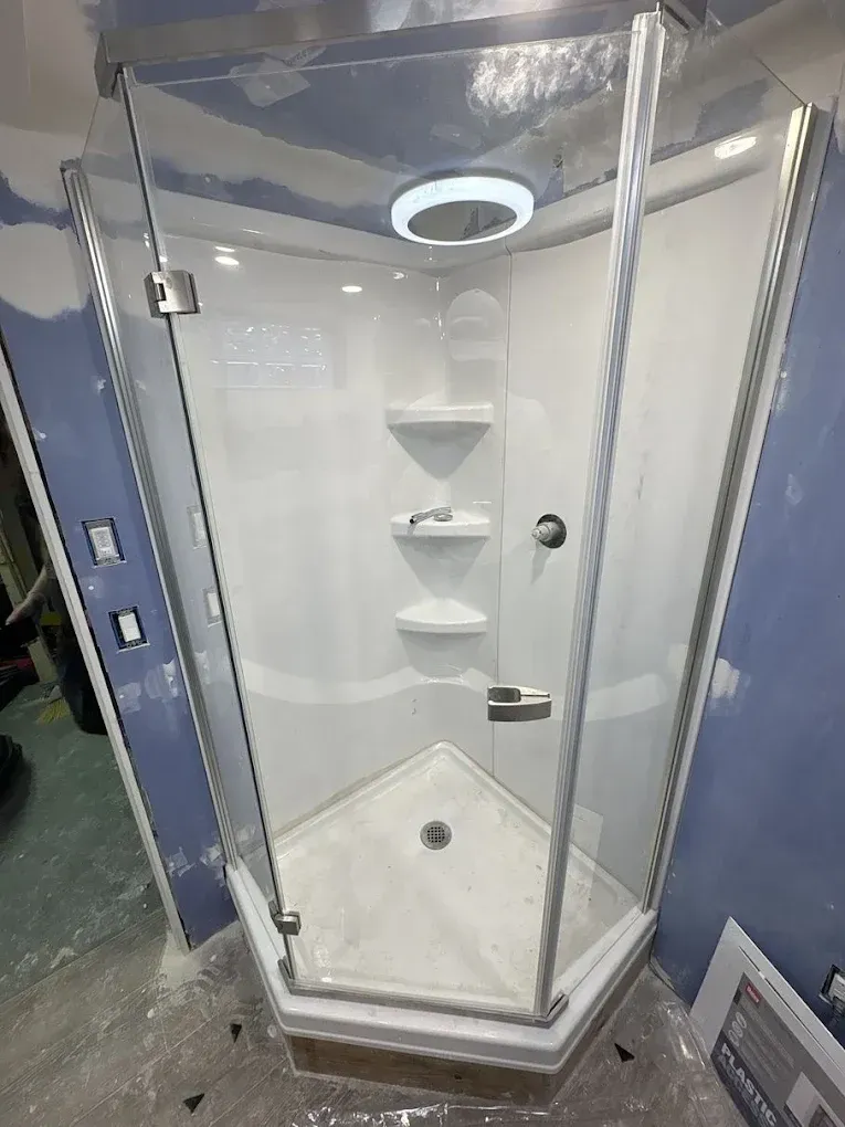 A white neo-angle shower stall with glass doors and built-in shelving, installed in a room with unfinished blue walls.