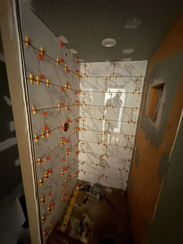 A tiled shower stall under construction with white marble-patterned tiles held in place by orange leveling clips.