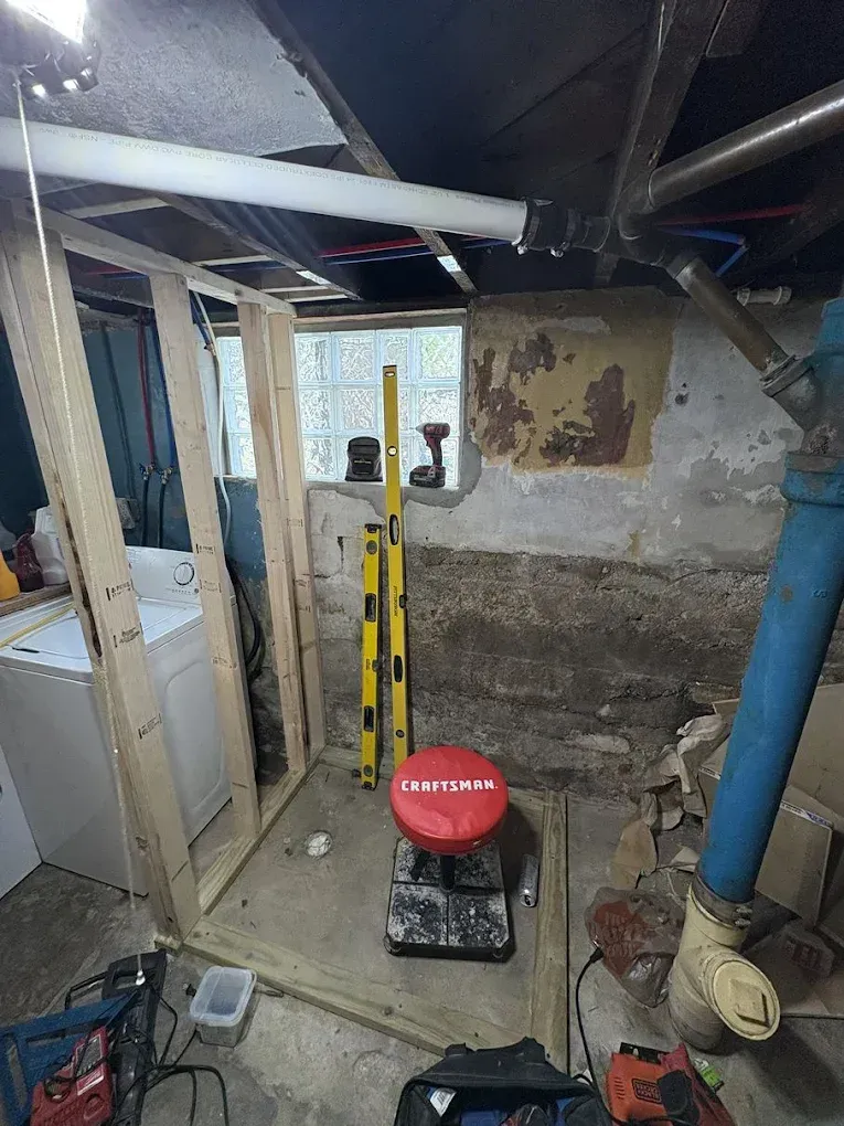 A partially framed wooden wall stands in a concrete basement near a washer, level tools, and a red stool.
