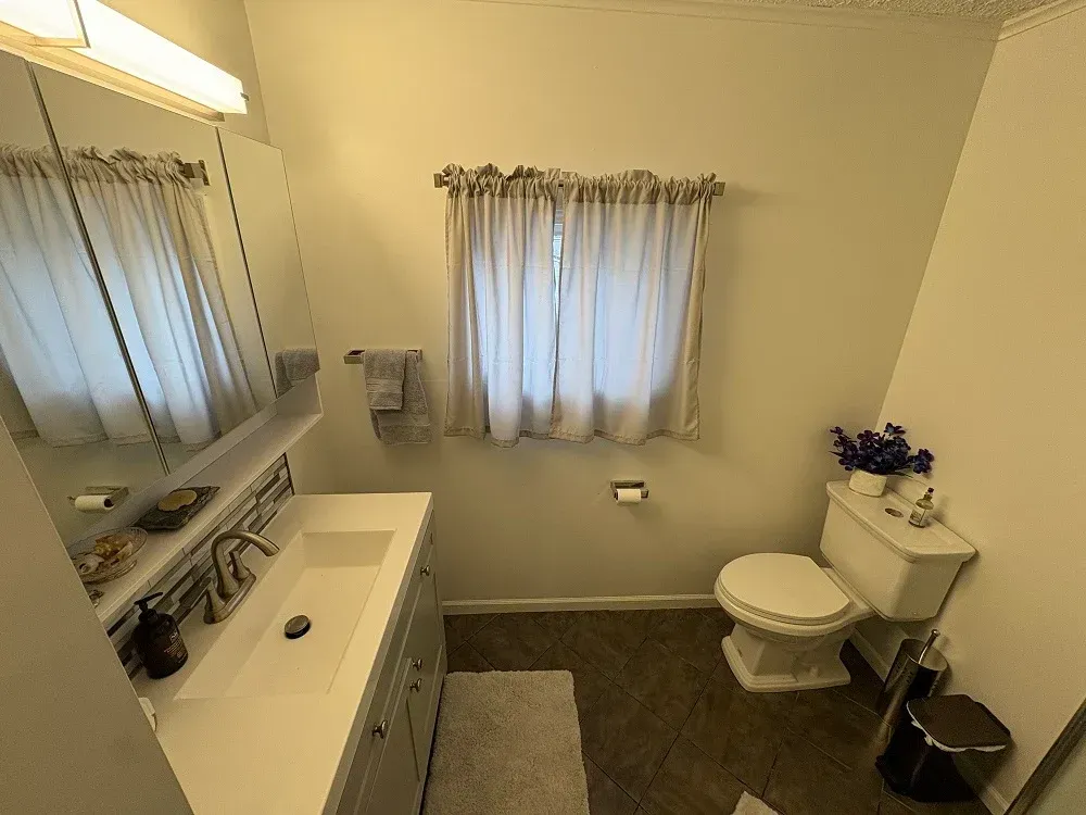 A bathroom featuring a white vanity with a sink, a large mirror, a window with beige curtains, and a white toilet.