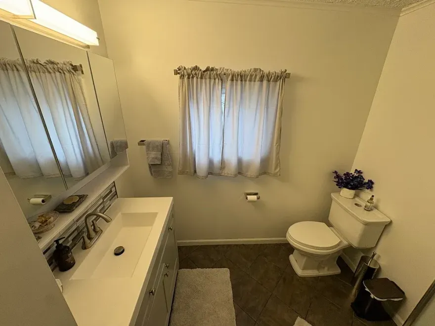 A well-lit, neutral-toned bathroom featuring a white vanity with a mirror, a toilet, and a window with light curtains.