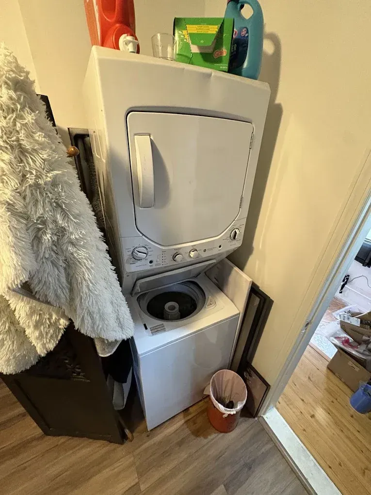 A white stacked laundry unit in a small alcove, with laundry supplies on top and a wastebasket on the floor.