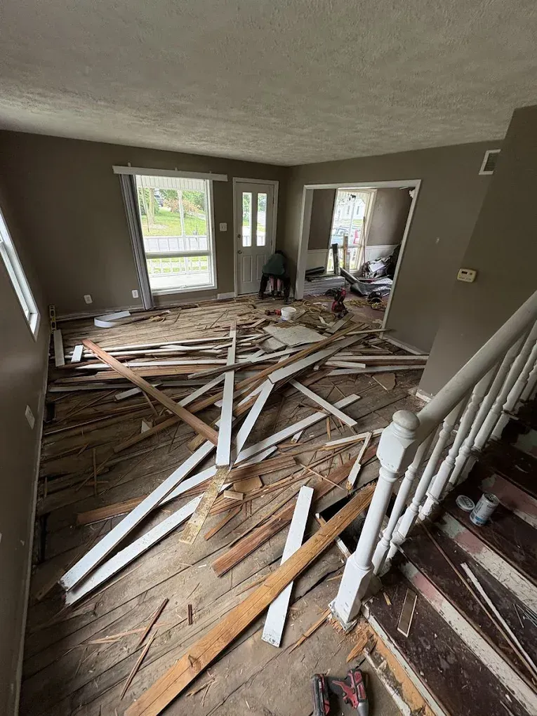 A room undergoing renovation with exposed wooden floorboards, scattered construction debris, and a wooden staircase.