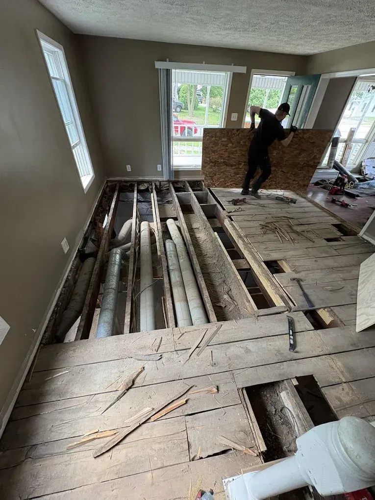 A person carries a plywood sheet over floor joists and exposed ductwork in a room undergoing subfloor renovation.
