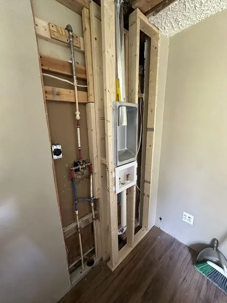 An open wall reveal showing laundry plumbing and a dryer vent box installed between wooden studs.