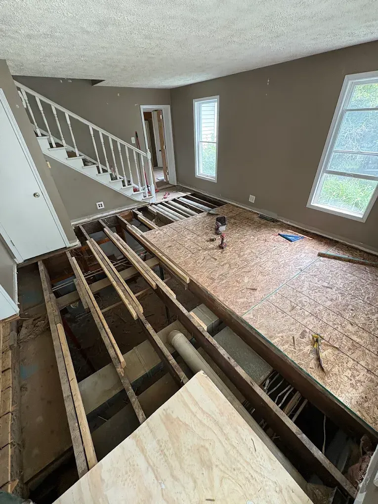 A living room under renovation with exposed floor joists next to a partially sub-floored area and a staircase.