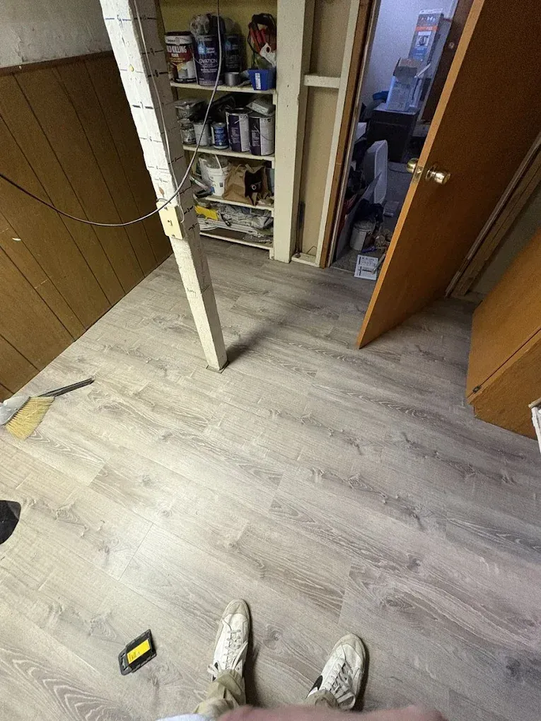 A first-person view of light wood-look vinyl flooring in a basement, with a support post, storage shelves, and an open door.