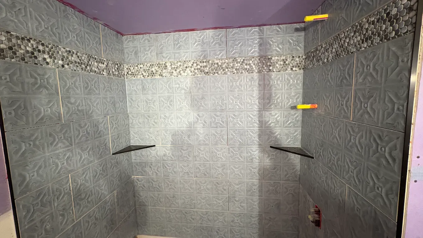 A shower stall with gray patterned tiles, a decorative mosaic accent strip, and two built-in corner shelves.