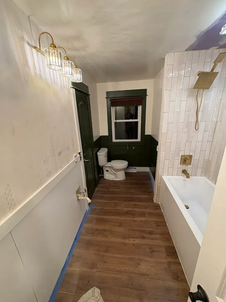 A bathroom under construction with wood-look flooring, a white bathtub with tiled walls, and a toilet by a small window.