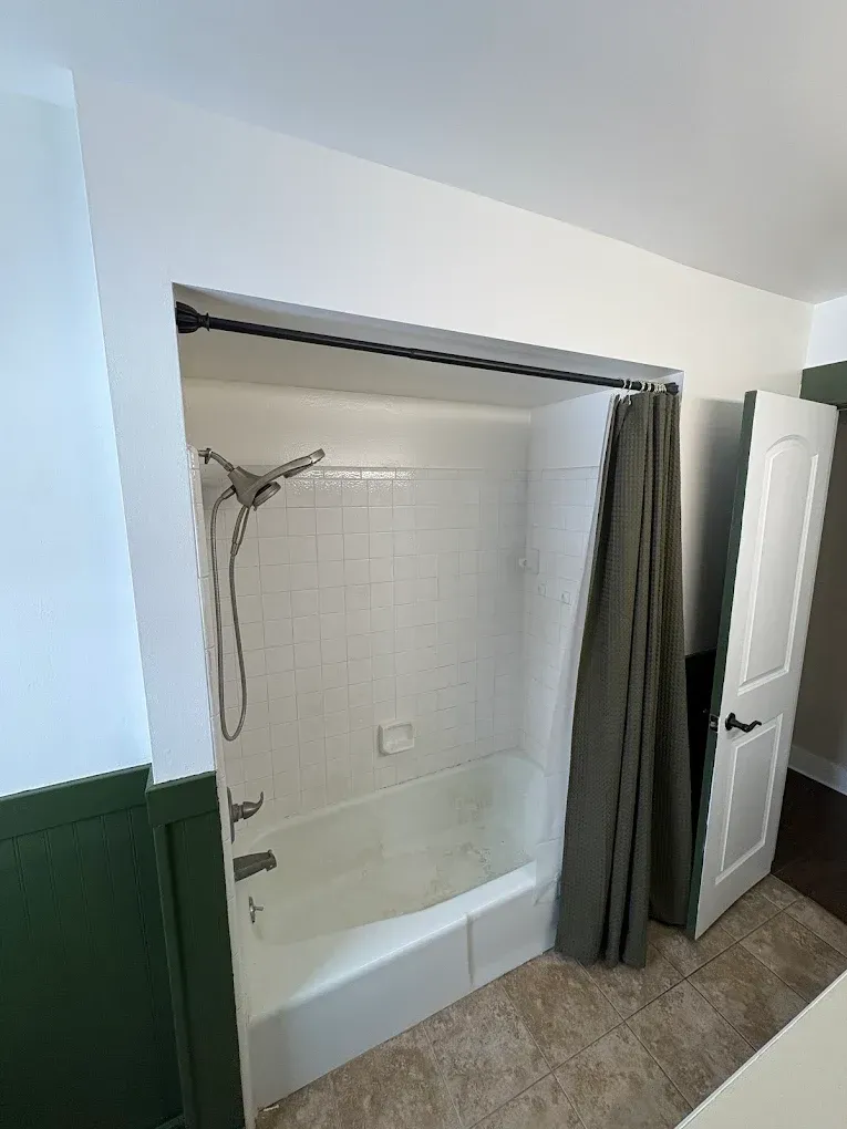 A white bathtub and shower stall with white tile walls, a handheld showerhead, and an olive green shower curtain.