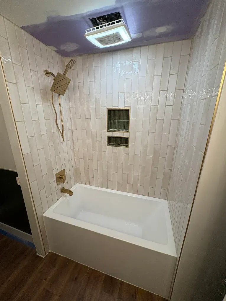 A white bathtub with gold fixtures installed in a shower stall featuring white vertical tiled walls and a recessed niche.